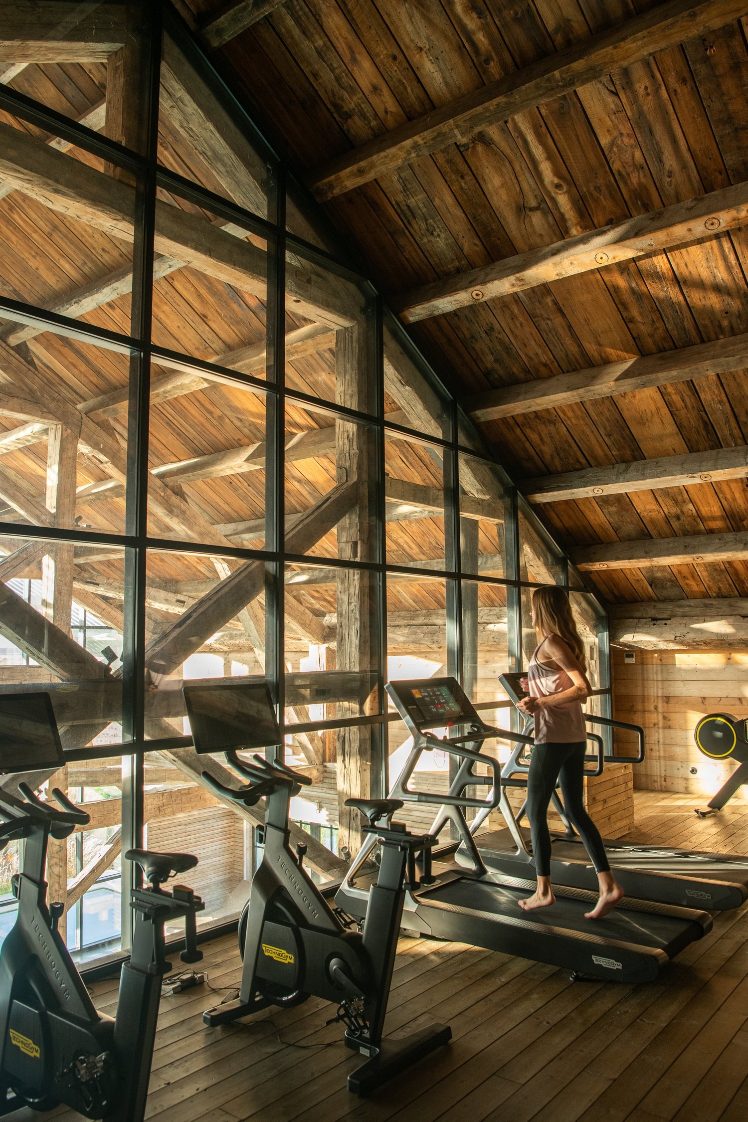 The gym set under a timber roof, treadmills and bikes facing a wall of glass that looks into the crossed beams of the old barn structure.