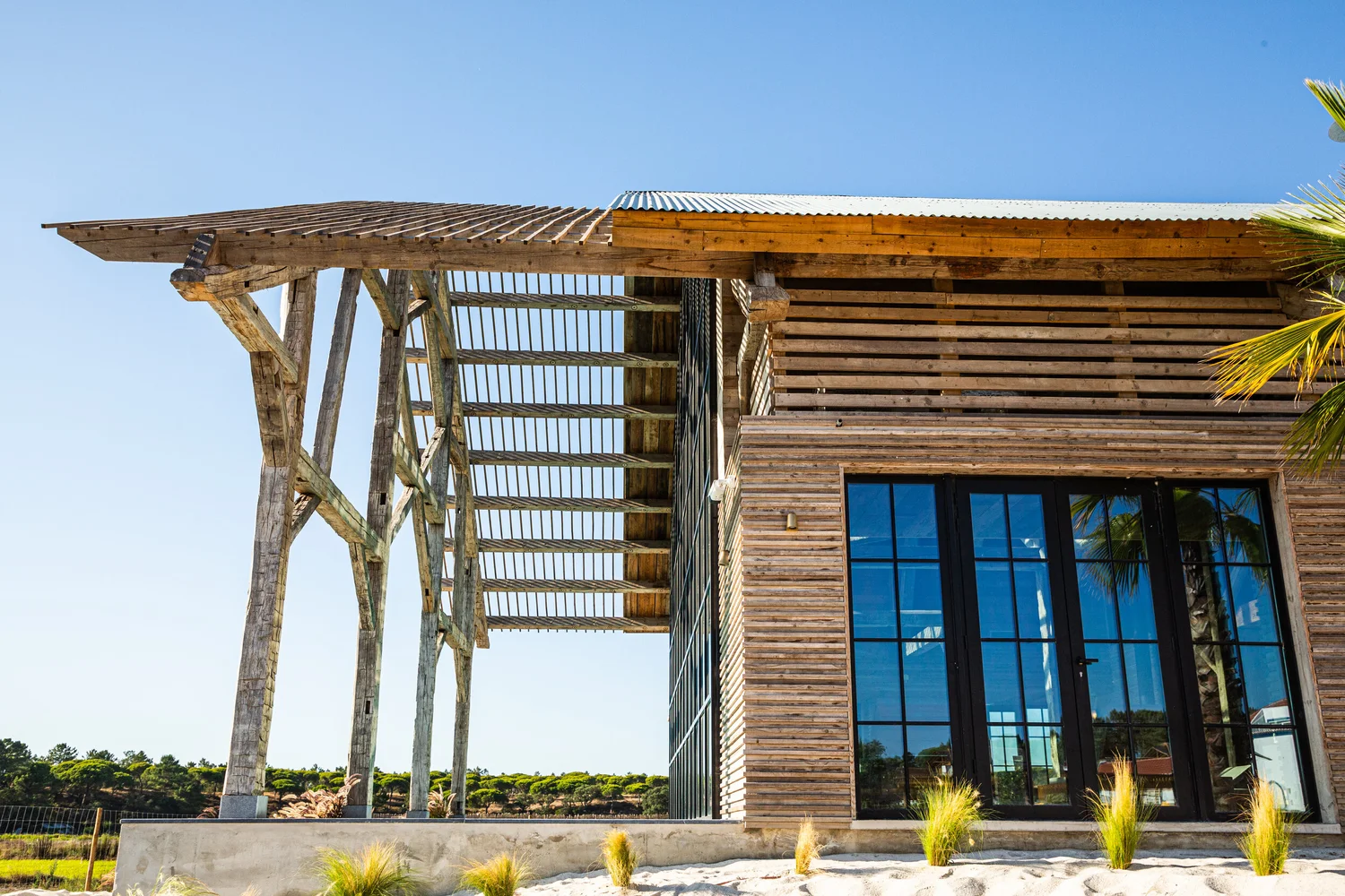 A weathered timber building with a corrugated roof and a long cantilevered brise-soleil on rough posts, tall black-framed windows below.