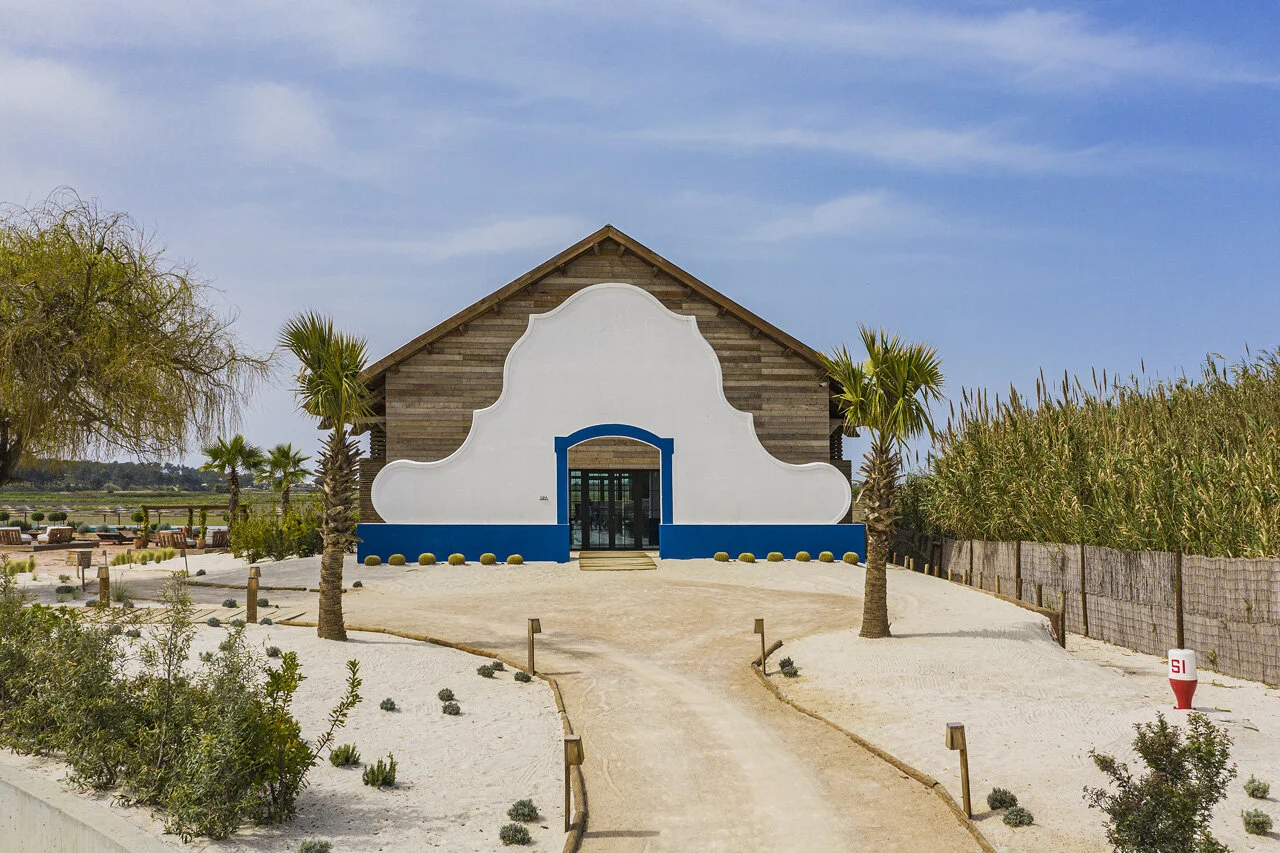 The gabled main building seen head on across raked sand, its white wall scalloped above a blue-painted plinth, palms either side and reeds behind.