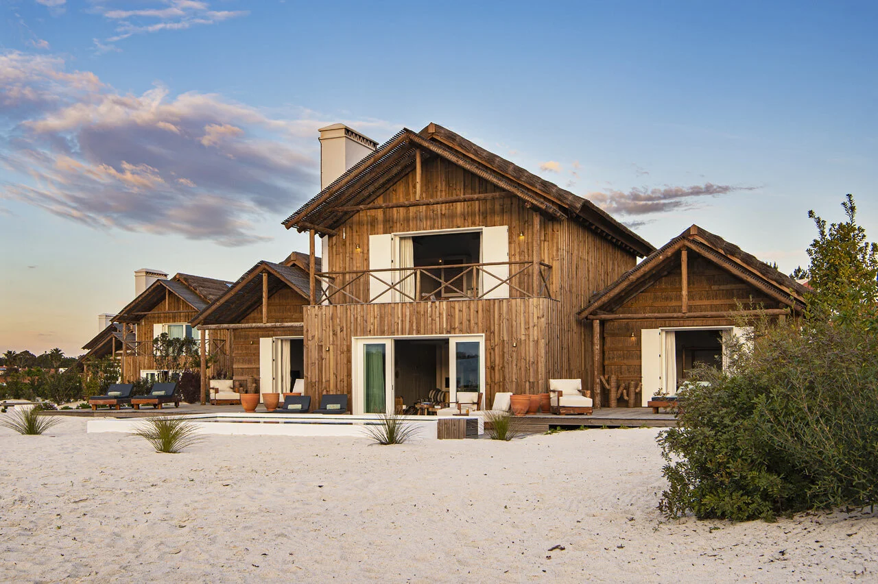 Timber-clad villas with steep gables standing on white sand at dusk, their doors folded open to a terrace and a small pool between them.