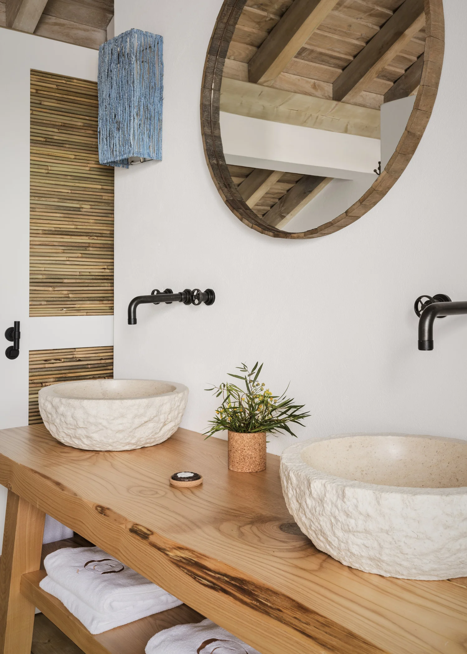 A bathroom vanity of thick raw timber with two rough-hewn stone basins, black wall taps and a round mirror reflecting the beamed ceiling.