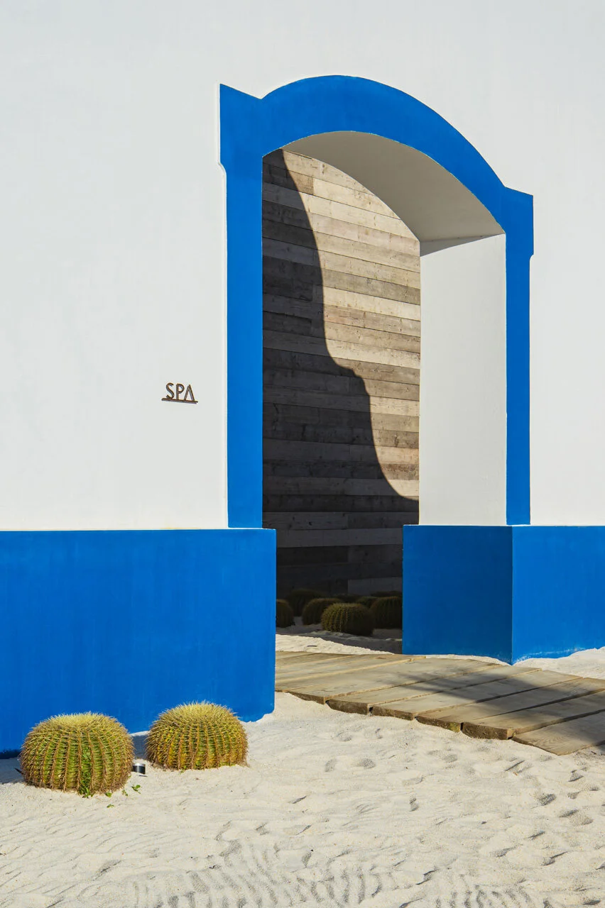 The spa entrance, a blue-painted arch cut in a white wall with the word SPA beside it, two barrel cacti on the sand and a timber ramp leading in.