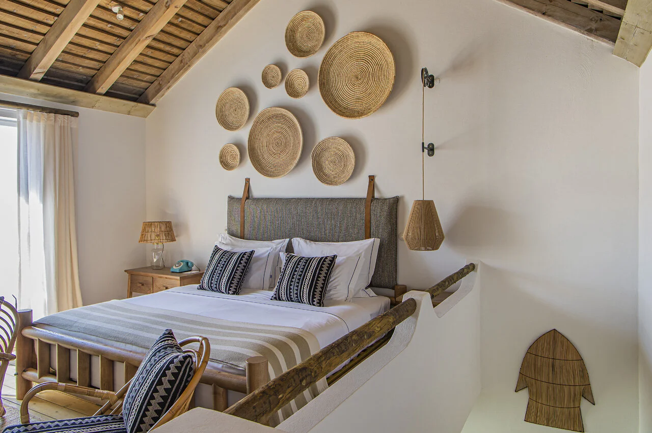 A bedroom under an exposed timber ceiling, woven discs arranged above the upholstered headboard and a rush pendant hanging beside the bed.