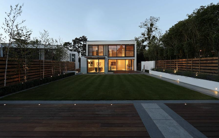 Modern two-storey house with large glass windows, illuminated interior, surrounded by a well-maintained lawn and outdoor lighting, with trees and fences on either side. Basement Render turning driveway