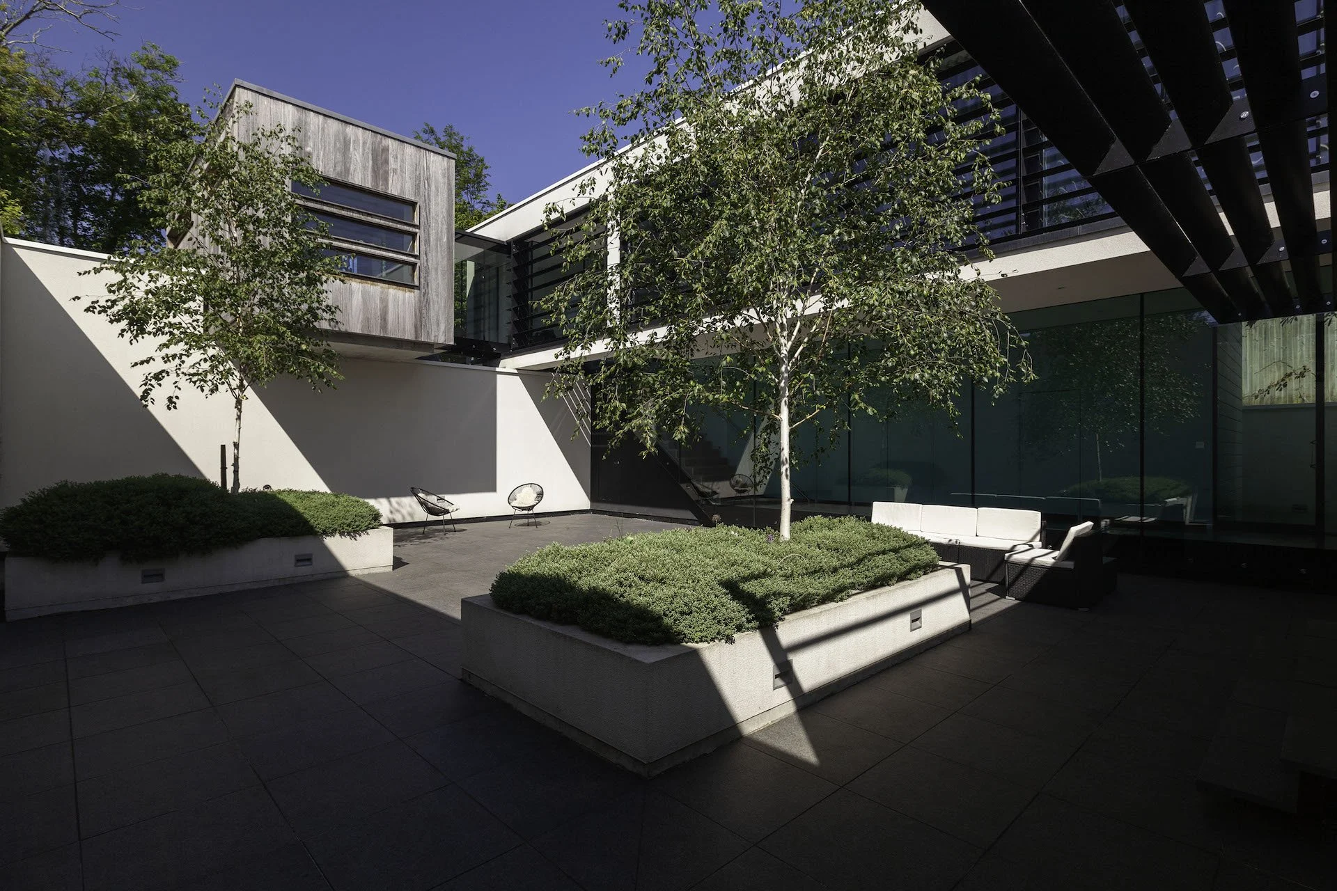 Modern outdoor courtyard with trees, greenery, and seating areas, surrounded by contemporary buildings with large glass windows and balconies.