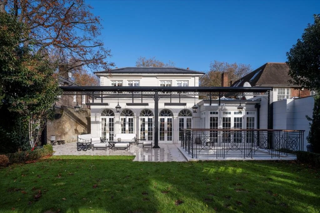 Backyard with a white two-story house, patio furniture, and a decorative metal railing, surrounded by green grass and trees, under a clear blue sky. refurbishment grade II listed building basement pool