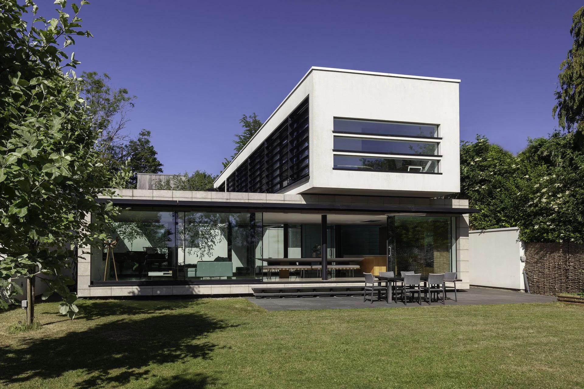 Modernist two-storey house with white exterior, large glass windows, and a balcony on the second floor, surrounded by trees and a well-maintained lawn.