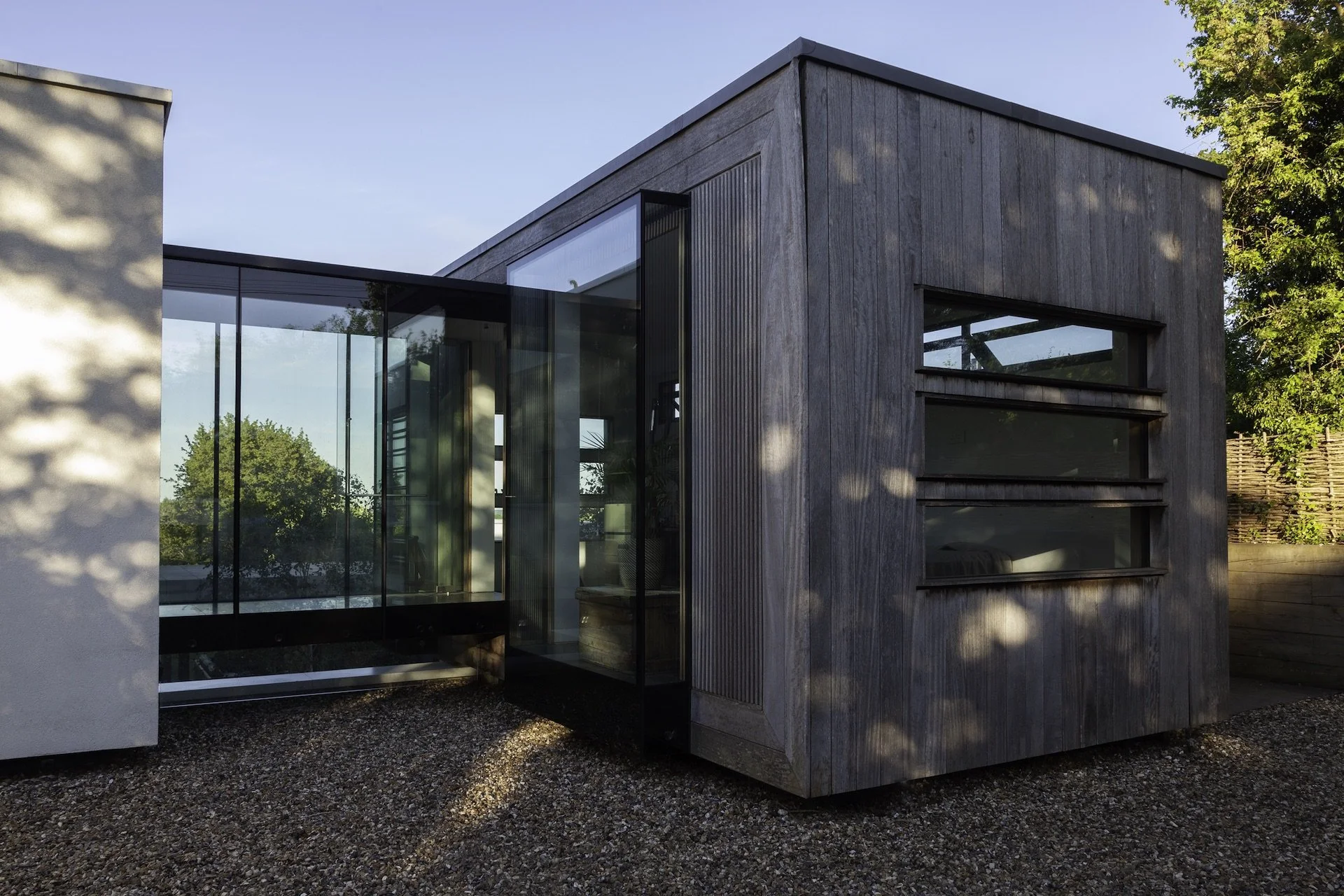 Modern house with wooden siding and large glass windows, surrounded by trees and a gravel ground.