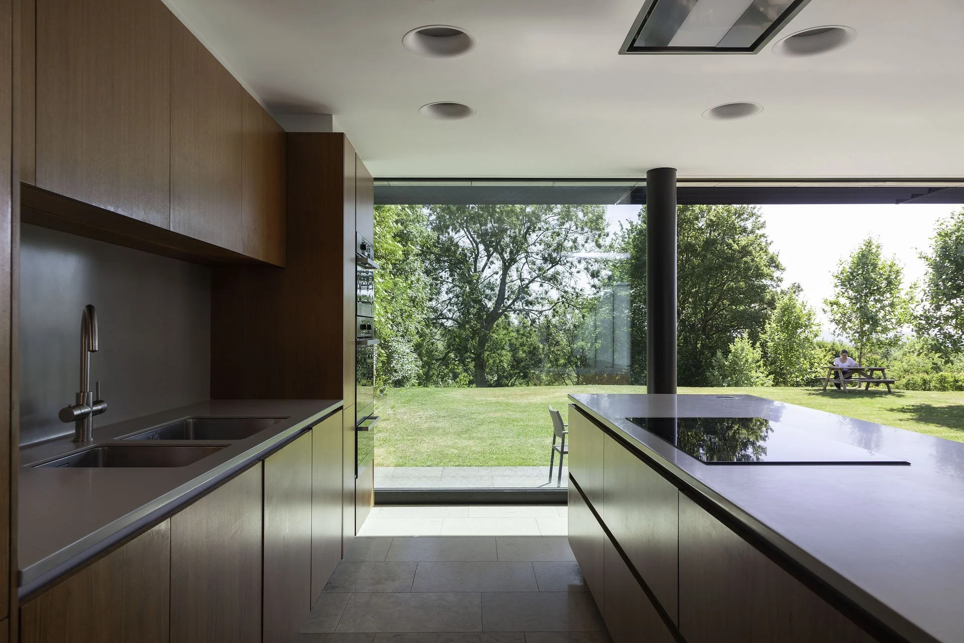 Modern kitchen with wooden cabinets, double sink, black stovetop, and large glass sliding door opening to a green outdoor landscape with trees and a person sitting at a picnic table.