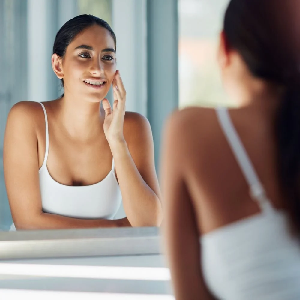 A woman with improved skin looking in the mirror after receiving a microneedling treatment.