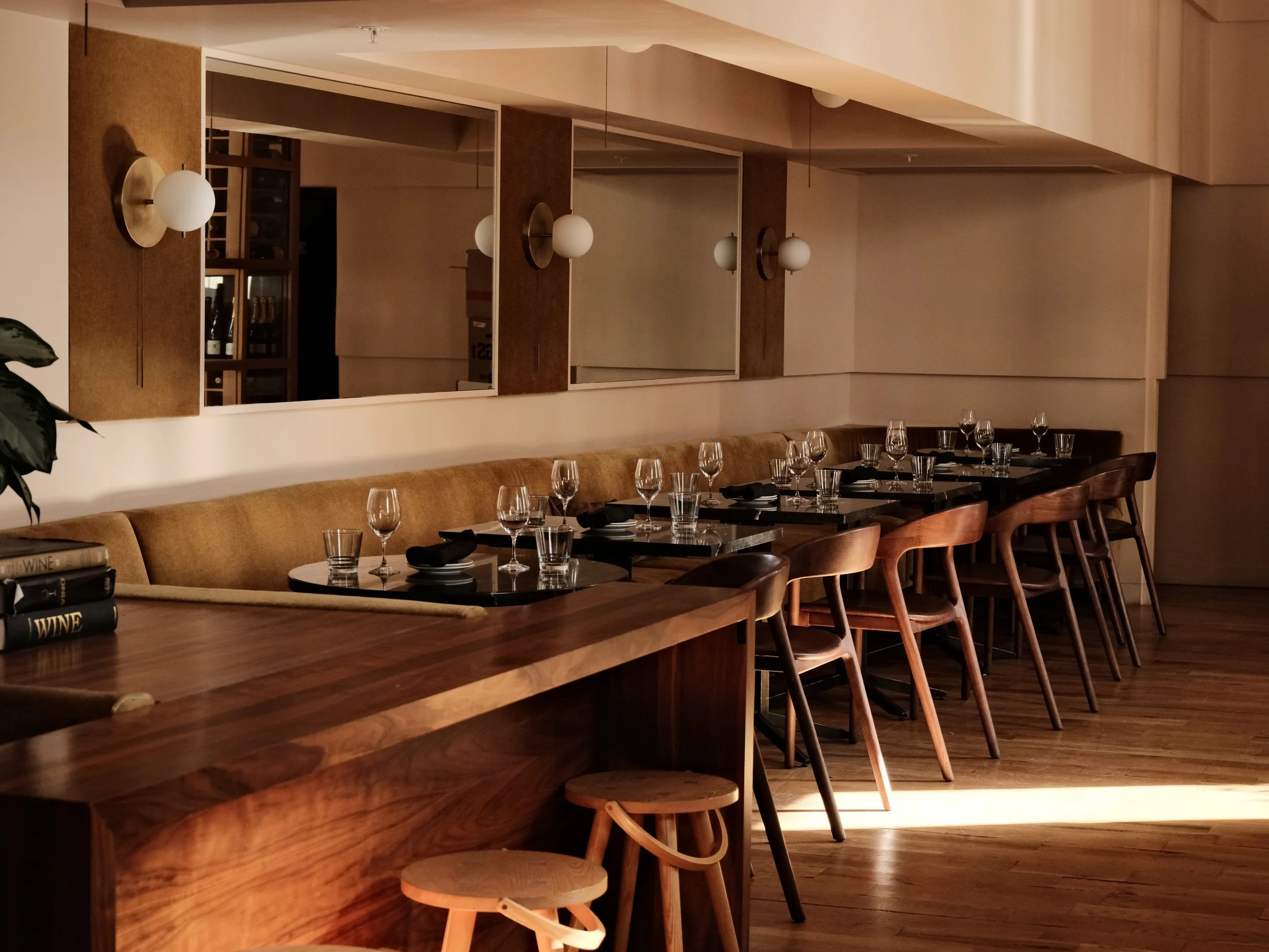 Empty restaurant dining area with a long wooden table, chairs, and wine glasses set for a meal, with warm lighting and a large mirror on the wall.