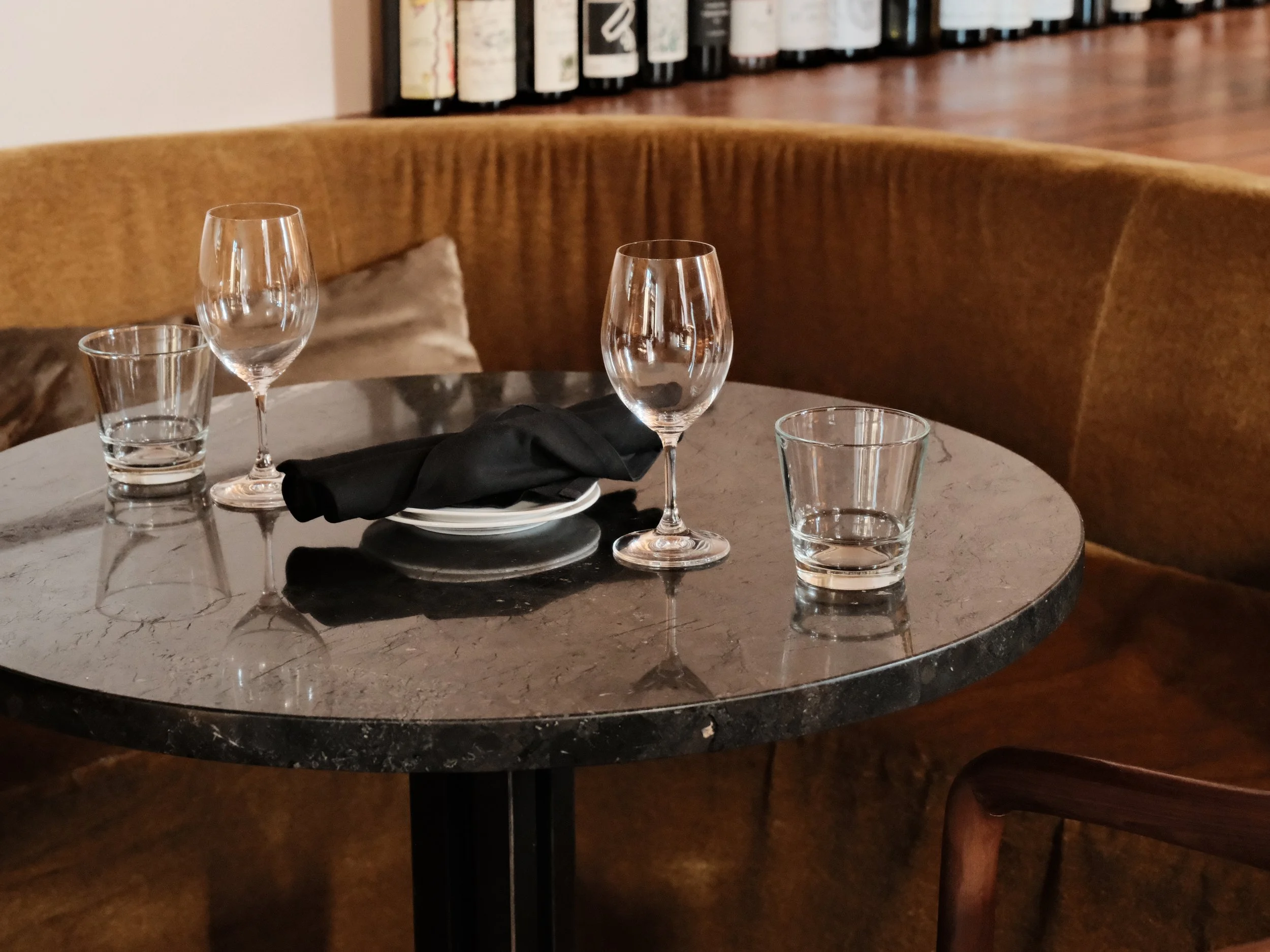 A round marble table set with two wine glasses, two water glasses, a black napkin on a small white plate, and a beige curved bench with a pillow in the background, in a restaurant or cafe setting.
