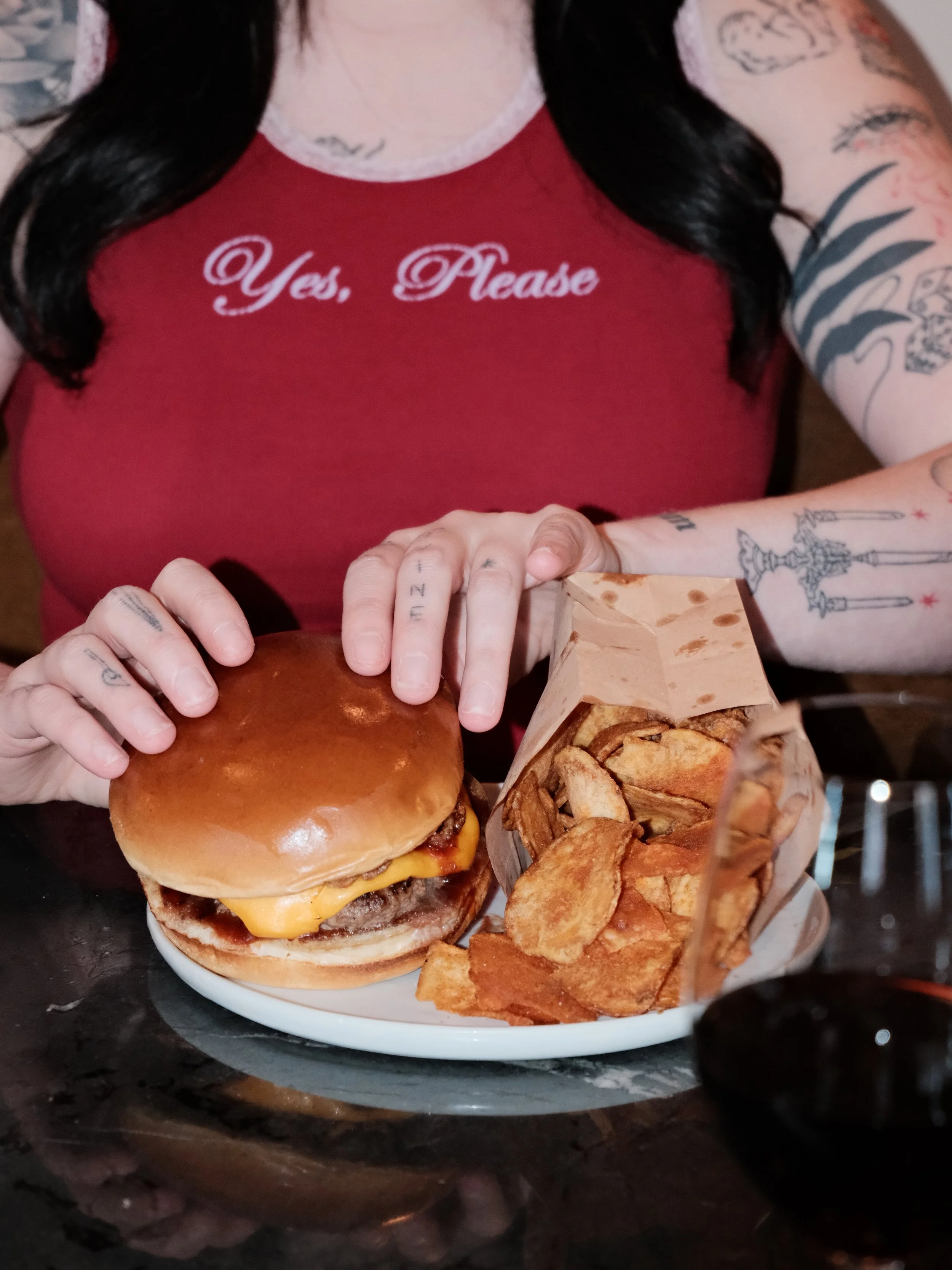 Person holding a cheeseburger with a side of potato chips and a drink on a table.