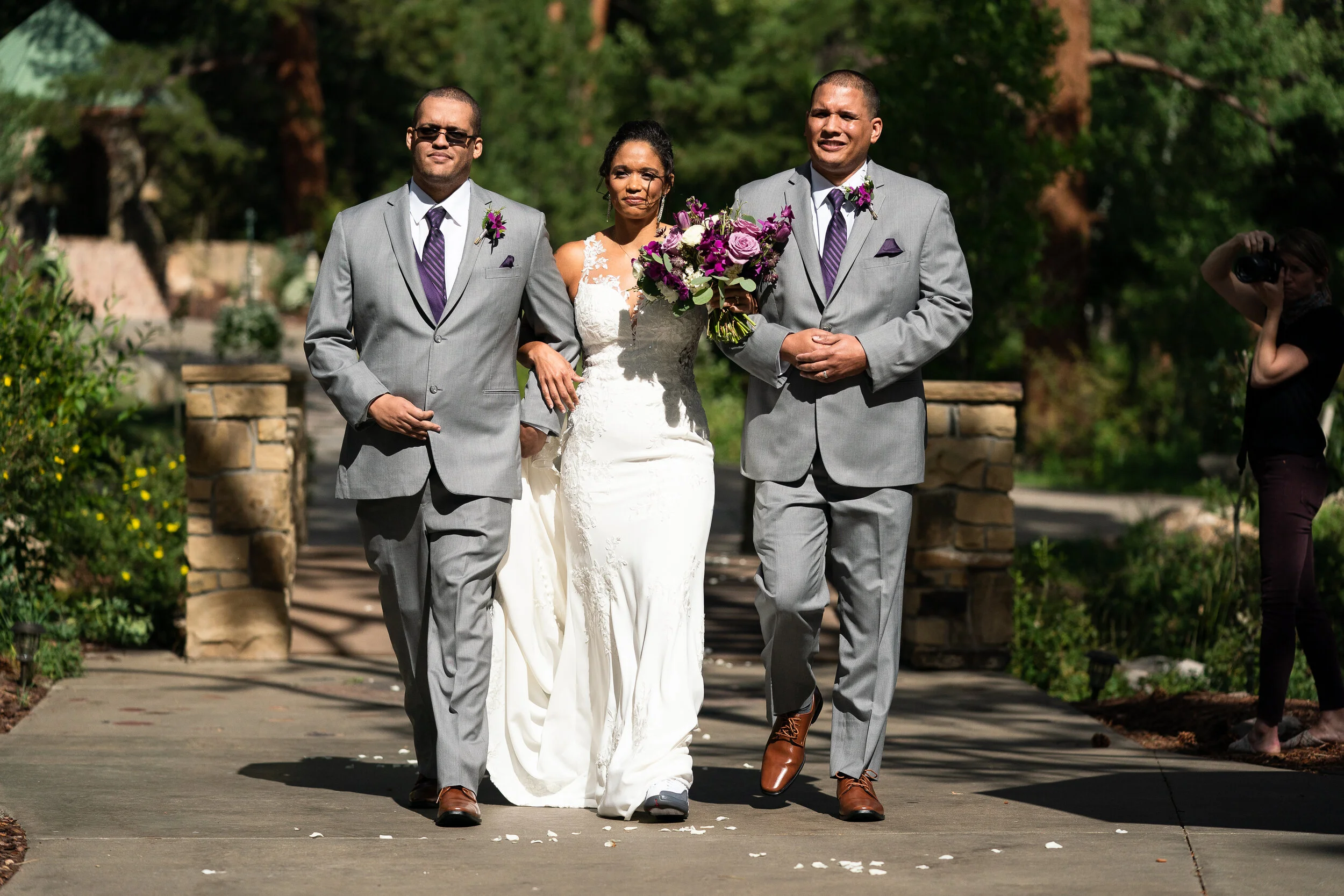 Bride with two men in gray suits walking down a path during an outdoor wedding ceremony, surrounded by greenery and a stone structure, with a photographer in the background.