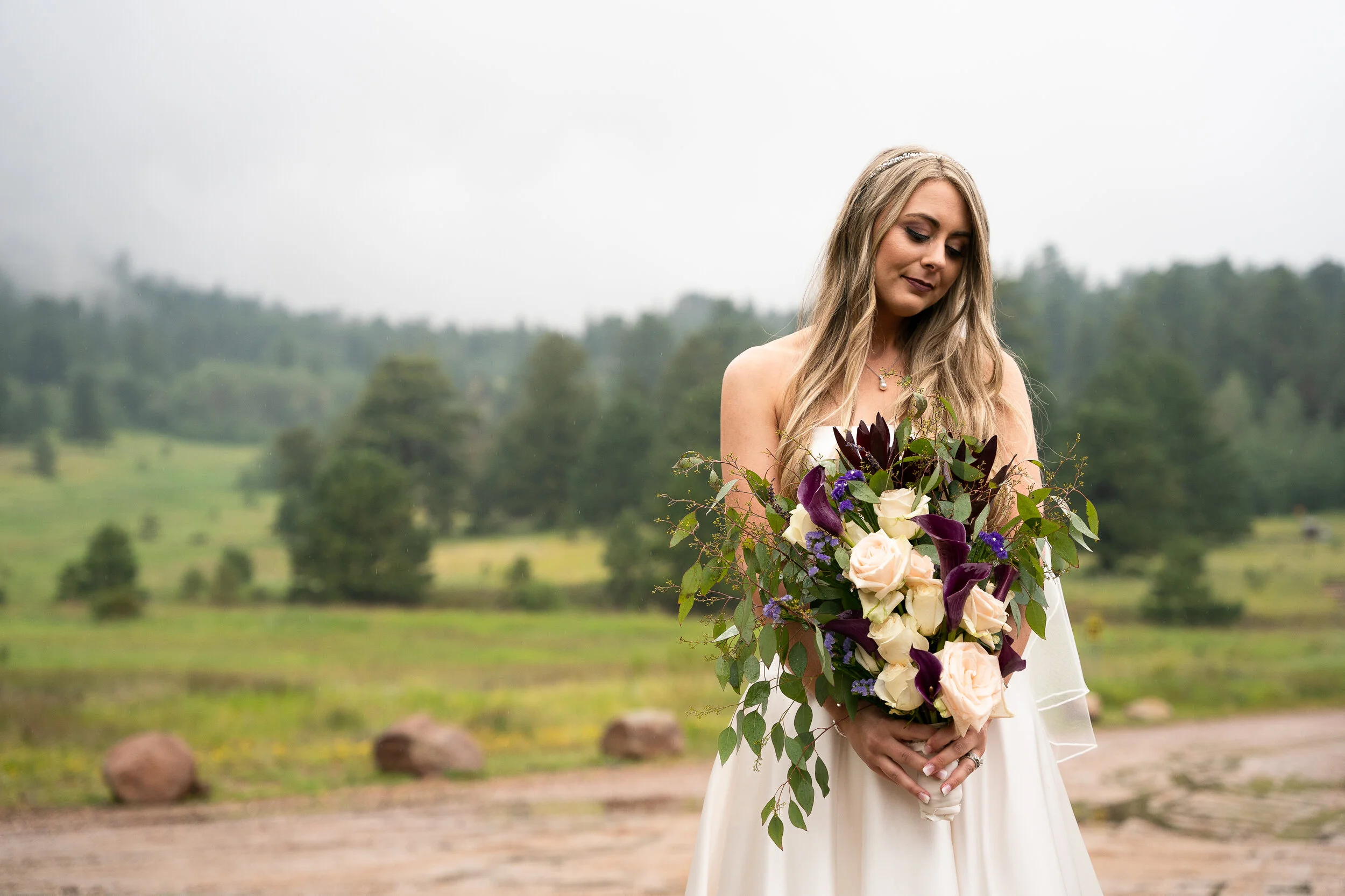 Bride holding a bouquet with roses and greenery, standing outdoors with forest background.