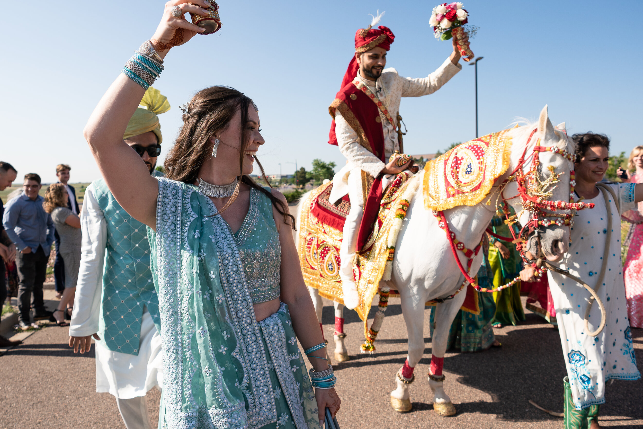 A traditional Indian wedding procession with a groom on a decorated white horse, a woman in a turquoise lehenga dancing, and people celebrating around them.