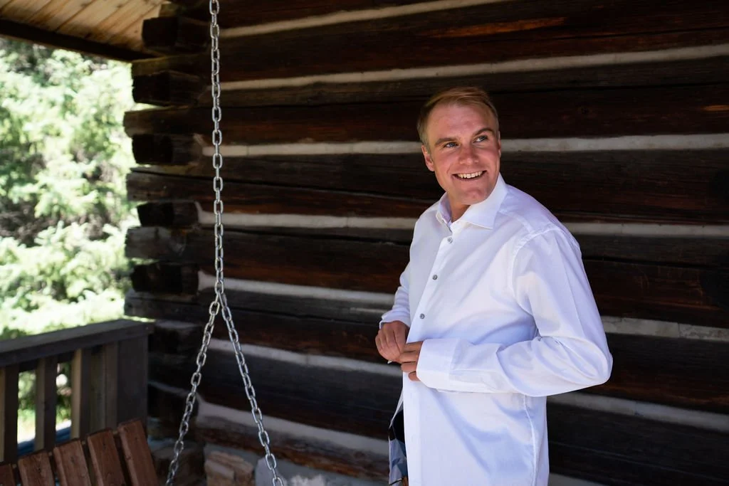 Smiling person in a white shirt stands on a porch with a wooden log wall in the background, next to a hanging wooden swing.