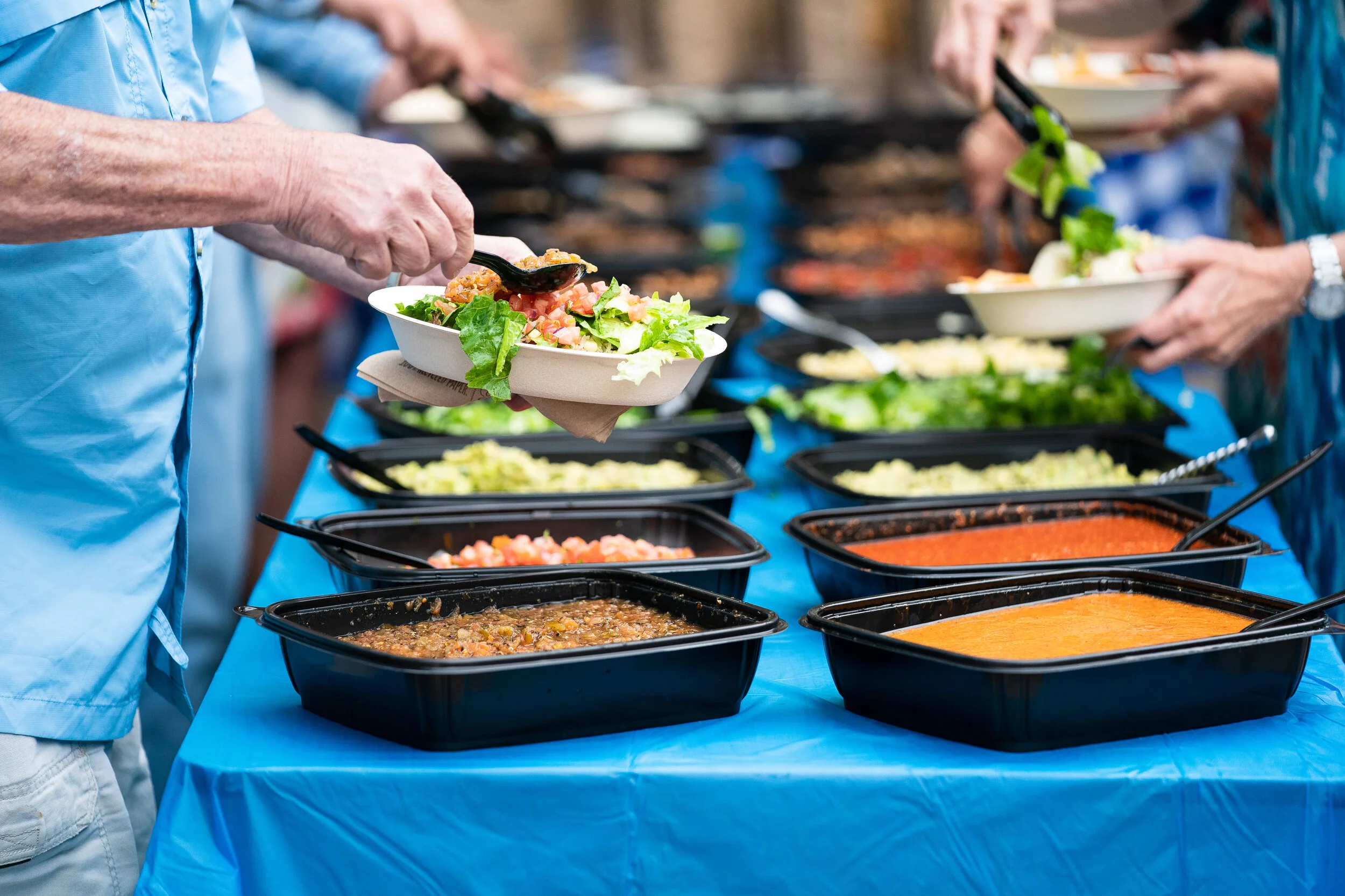 People serving themselves food from a buffet table with various dishes in black trays, including salads and sauces, on a blue tablecloth.