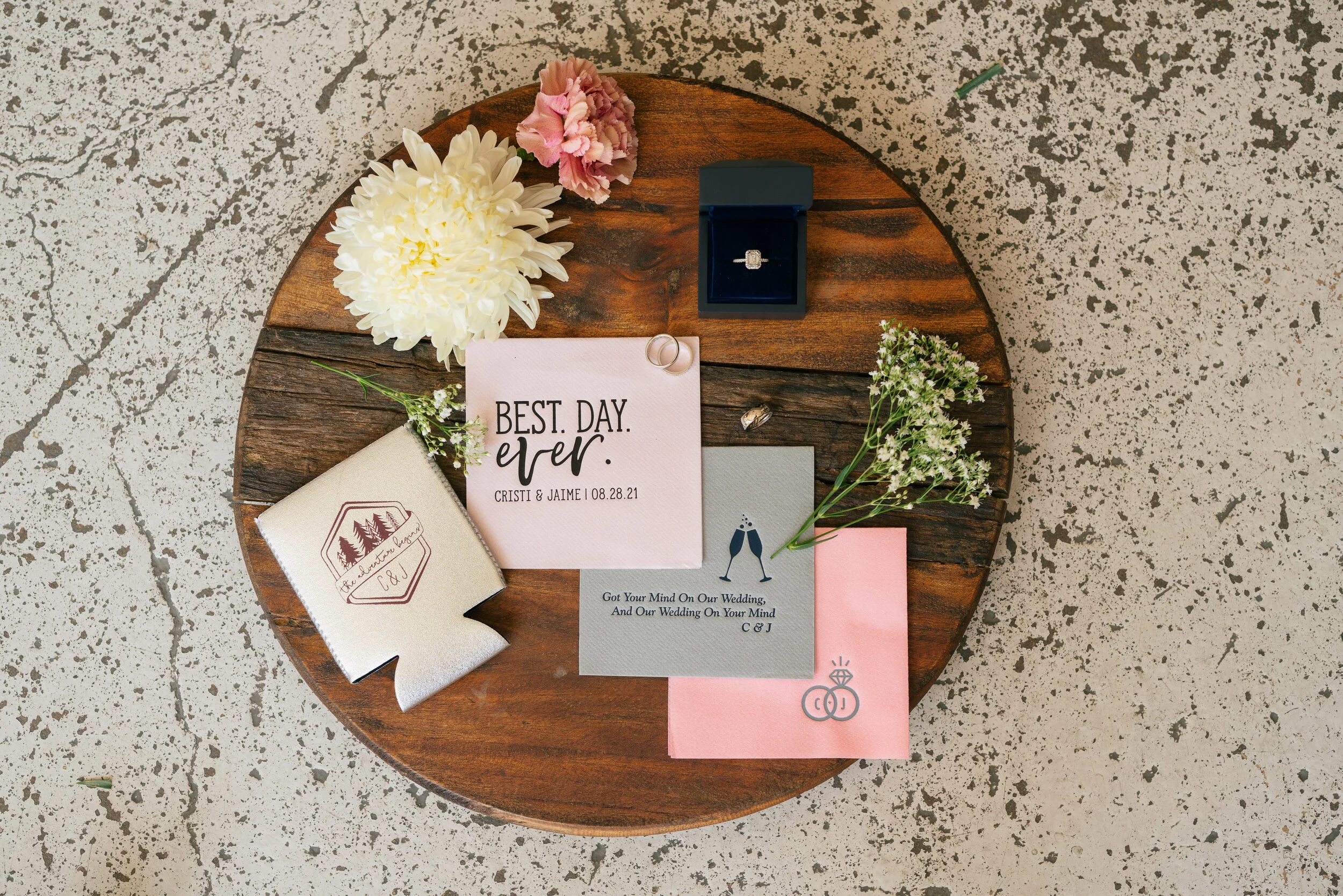 Flat lay of wedding-themed items on a wooden surface, including a 'Best Day Ever' card, wedding ring in a box, floral arrangements, and decorative napkins.