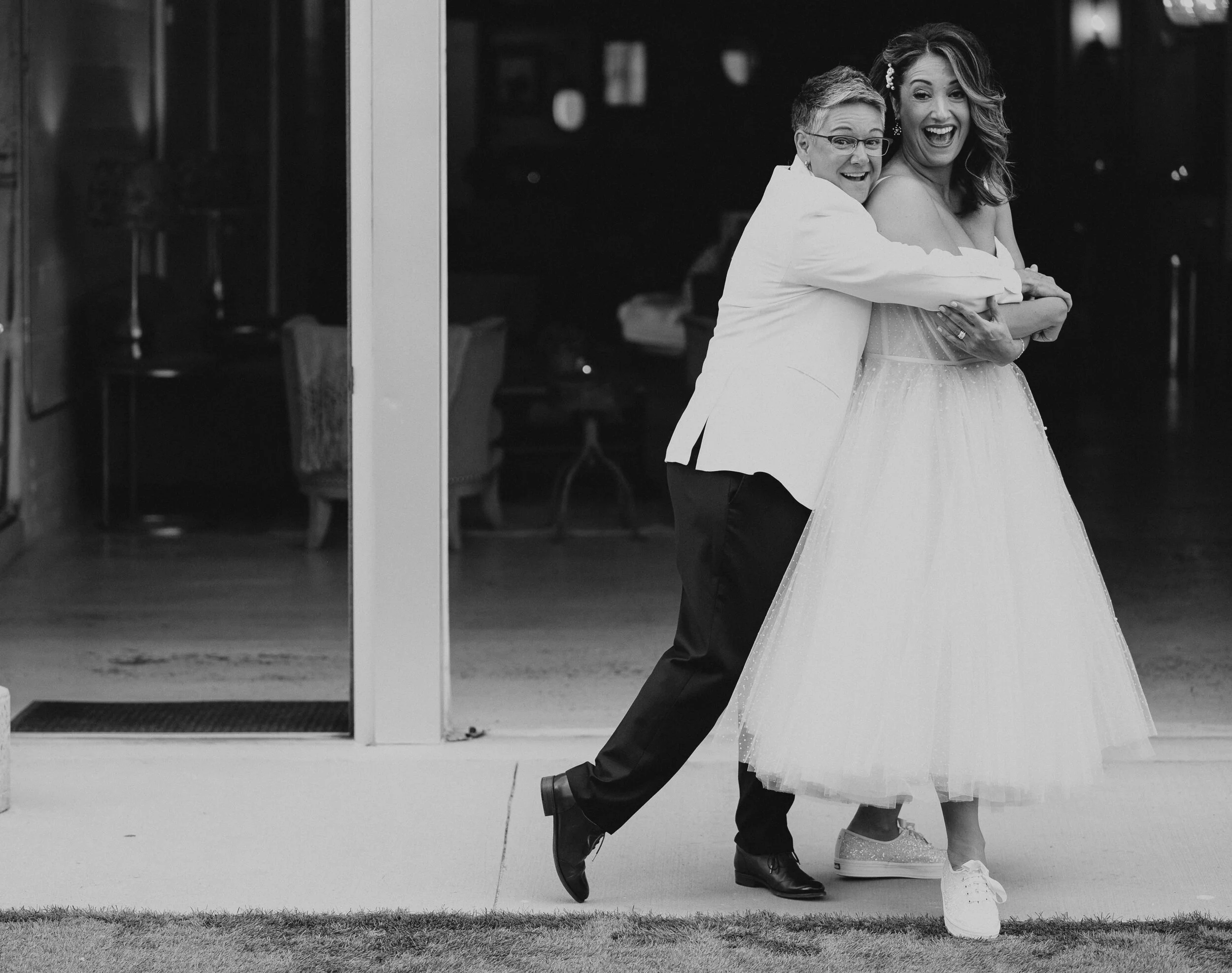 A joyful couple in formal attire, with one person in a white tuxedo jacket and another in a bridal gown, laughing and hugging in a playful pose outside a building.