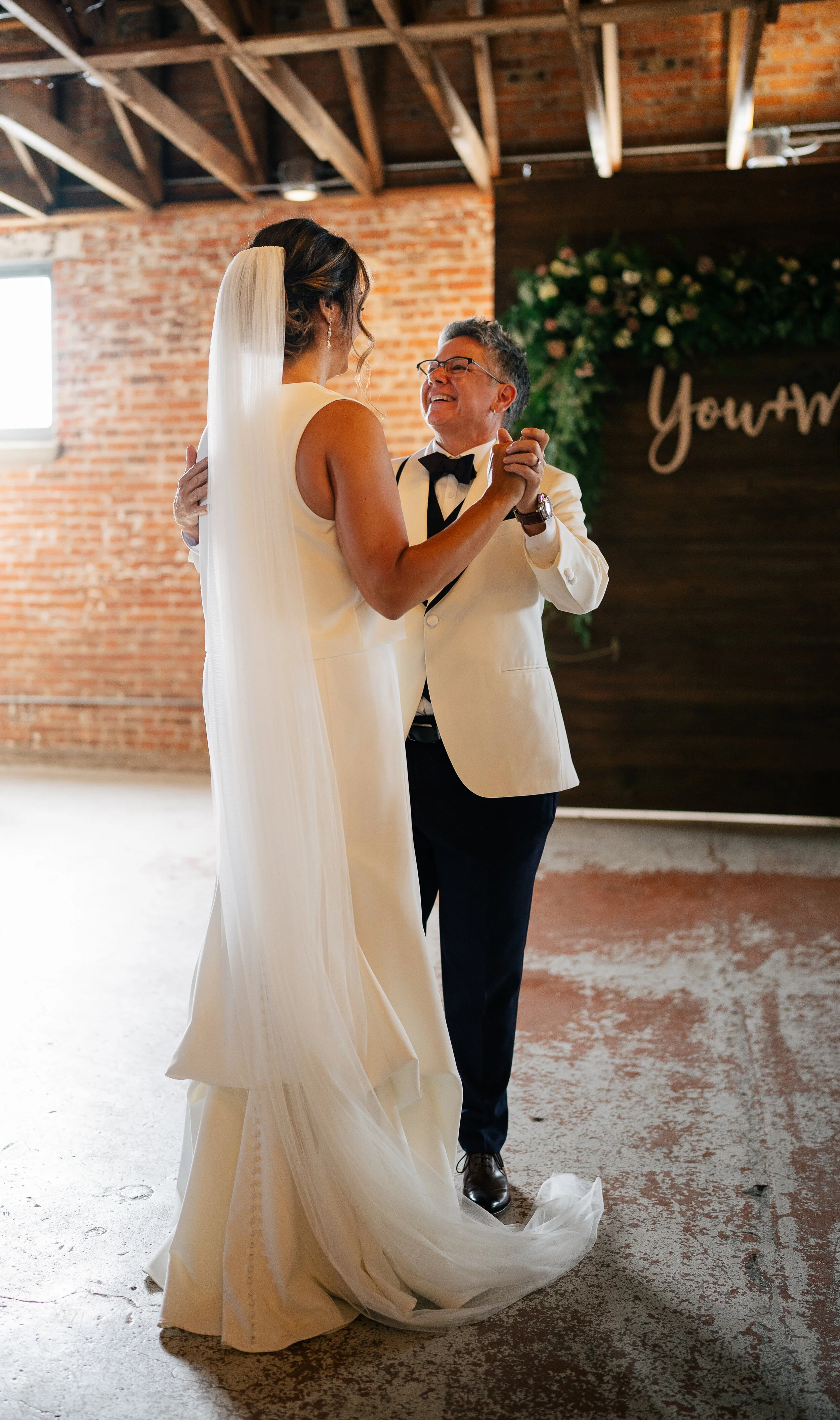 A couple dancing at a wedding, with one wearing a white dress and veil, and the other in a white suit jacket, against a rustic brick and wooden backdrop with greenery and a partial sign.