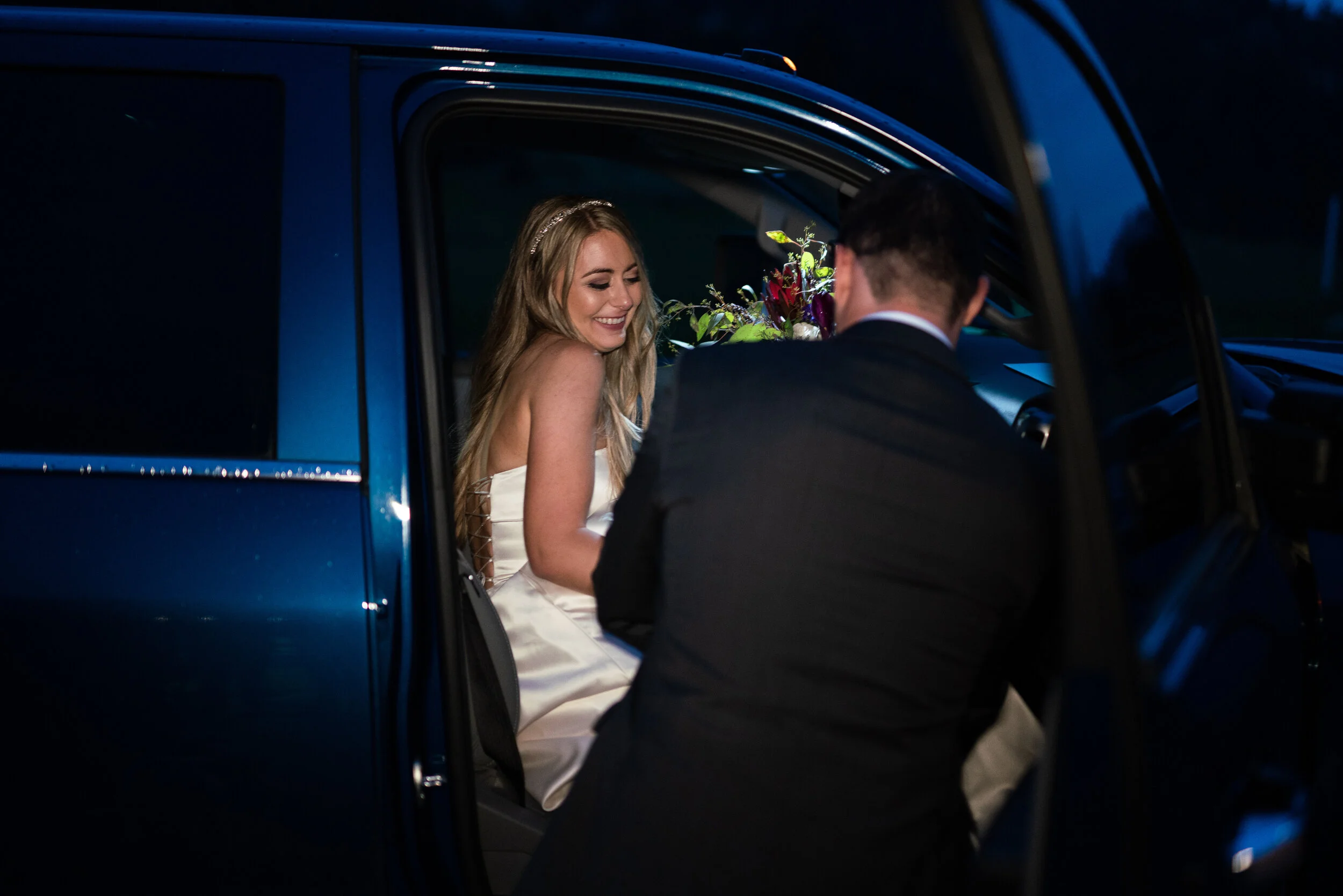 Bride sitting in a car, smiling, while a man in a suit stands outside holding a bouquet.