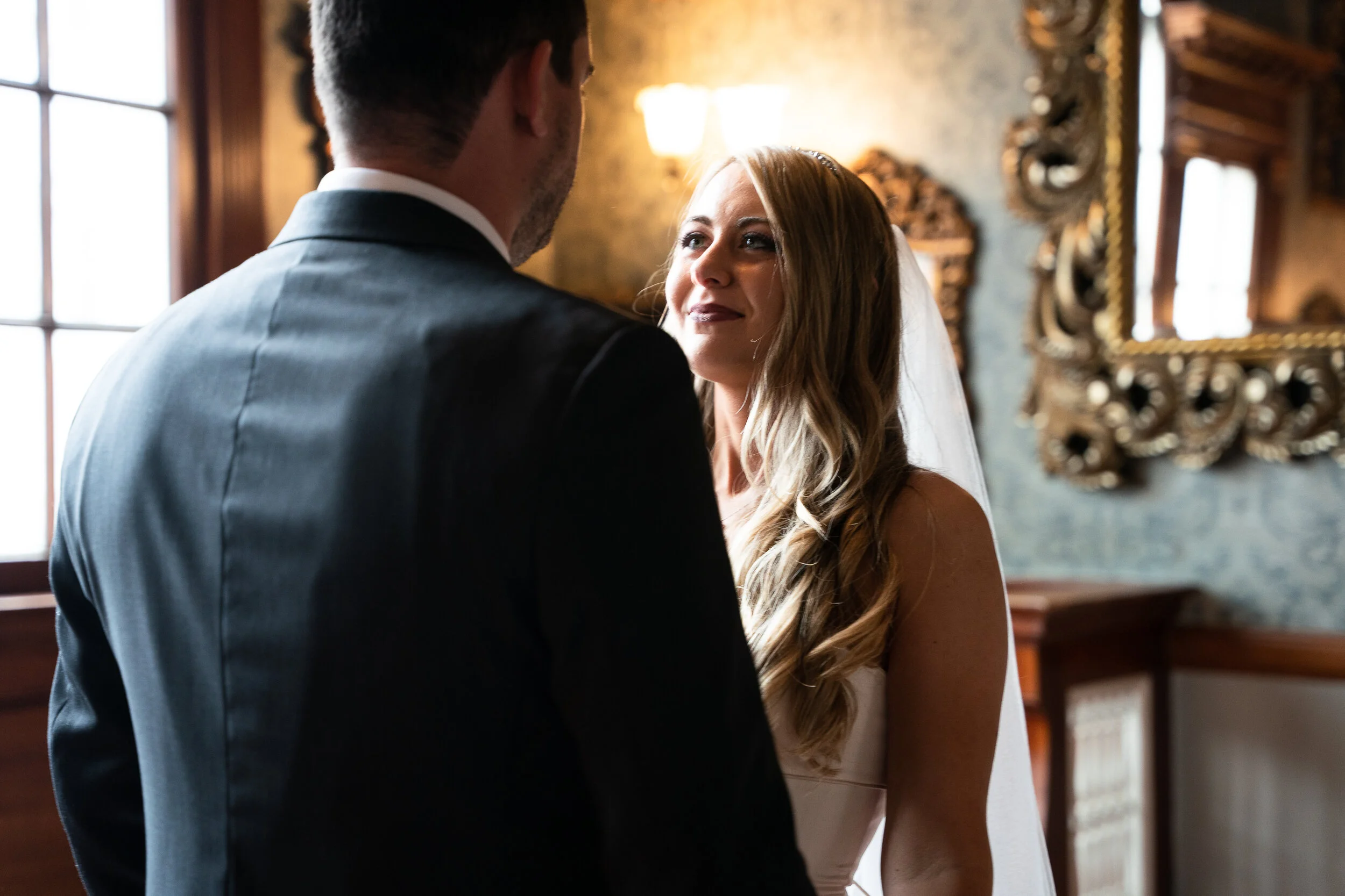Bride and groom exchanging vows indoors near a window, with elegant decor in the background.