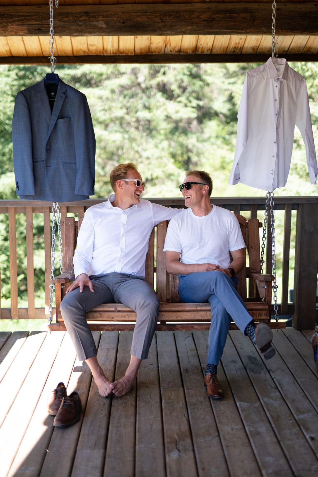 Two men sitting on a wooden swing under a porch, laughing and wearing sunglasses; a suit jacket and dress shirt hang on chains above them.