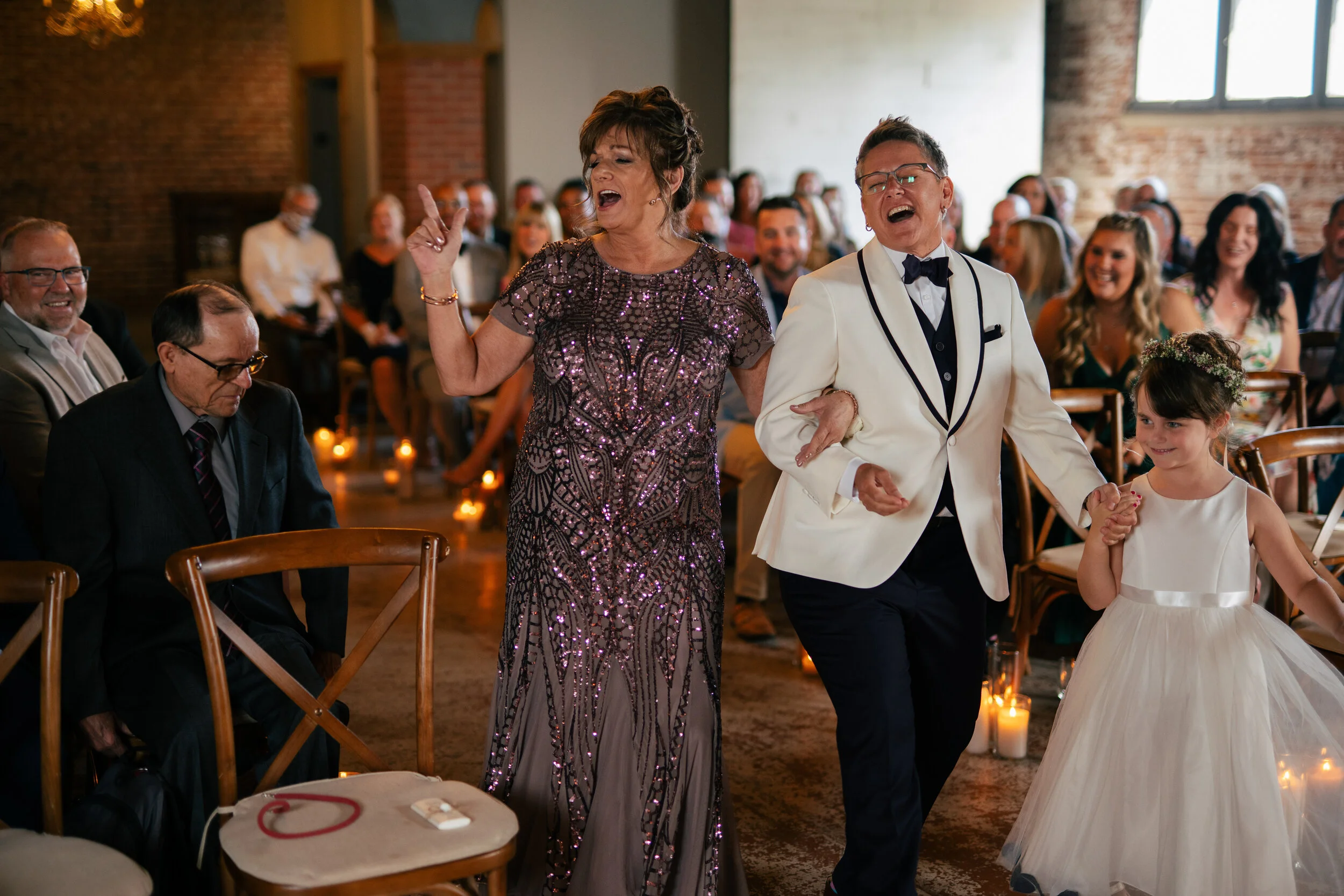 A joyful wedding procession with a woman in a sparkling dress, a person in a white tuxedo, and a girl in a white dress walking down an aisle lined with candles; smiling guests seated in the background.
