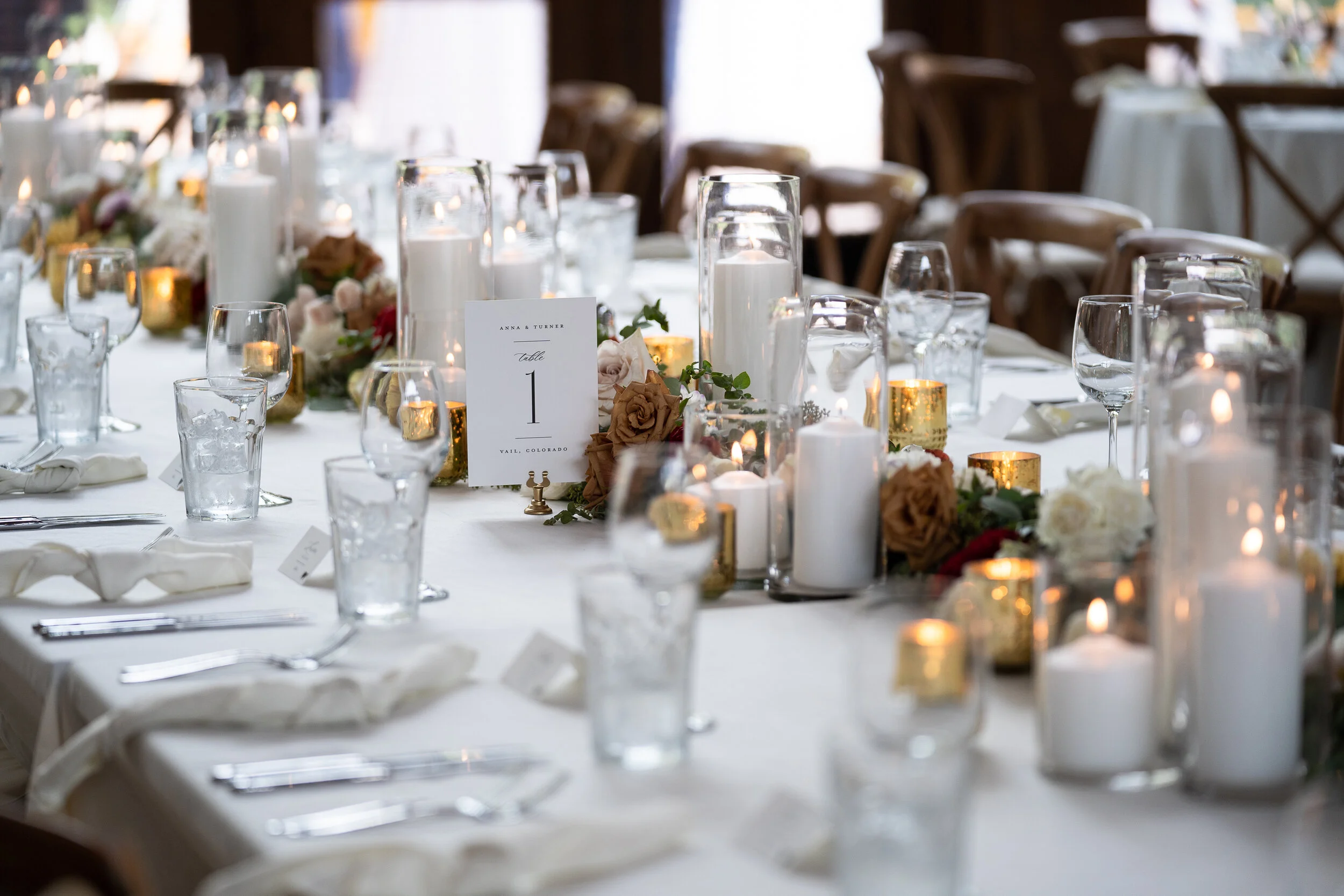 Elegant wedding reception table setting with white tablecloth, candles, and floral arrangements. "Table 1" card displayed, surrounded by glassware and cutlery.