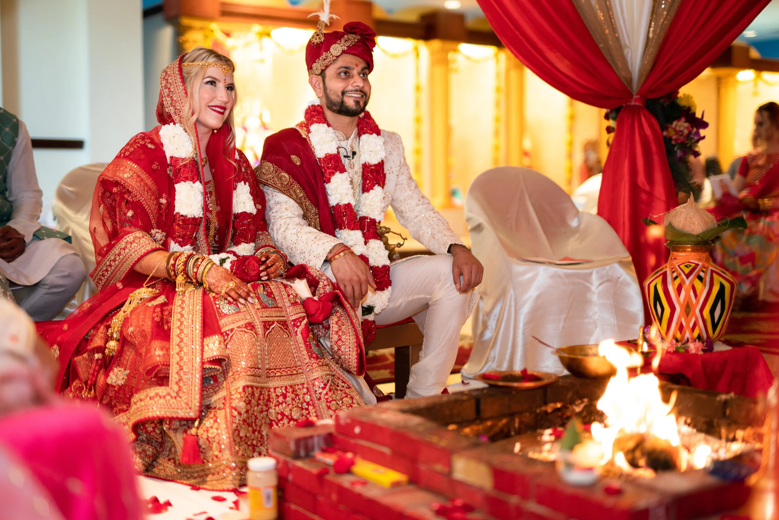A couple in traditional Indian wedding attire sitting near a ceremonial fire during a wedding ritual.