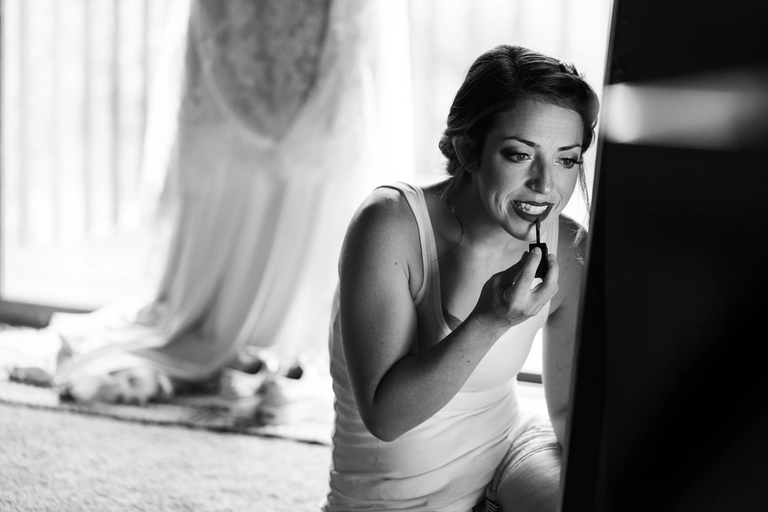 Woman applying lipstick in front of a mirror, wedding dress in background.