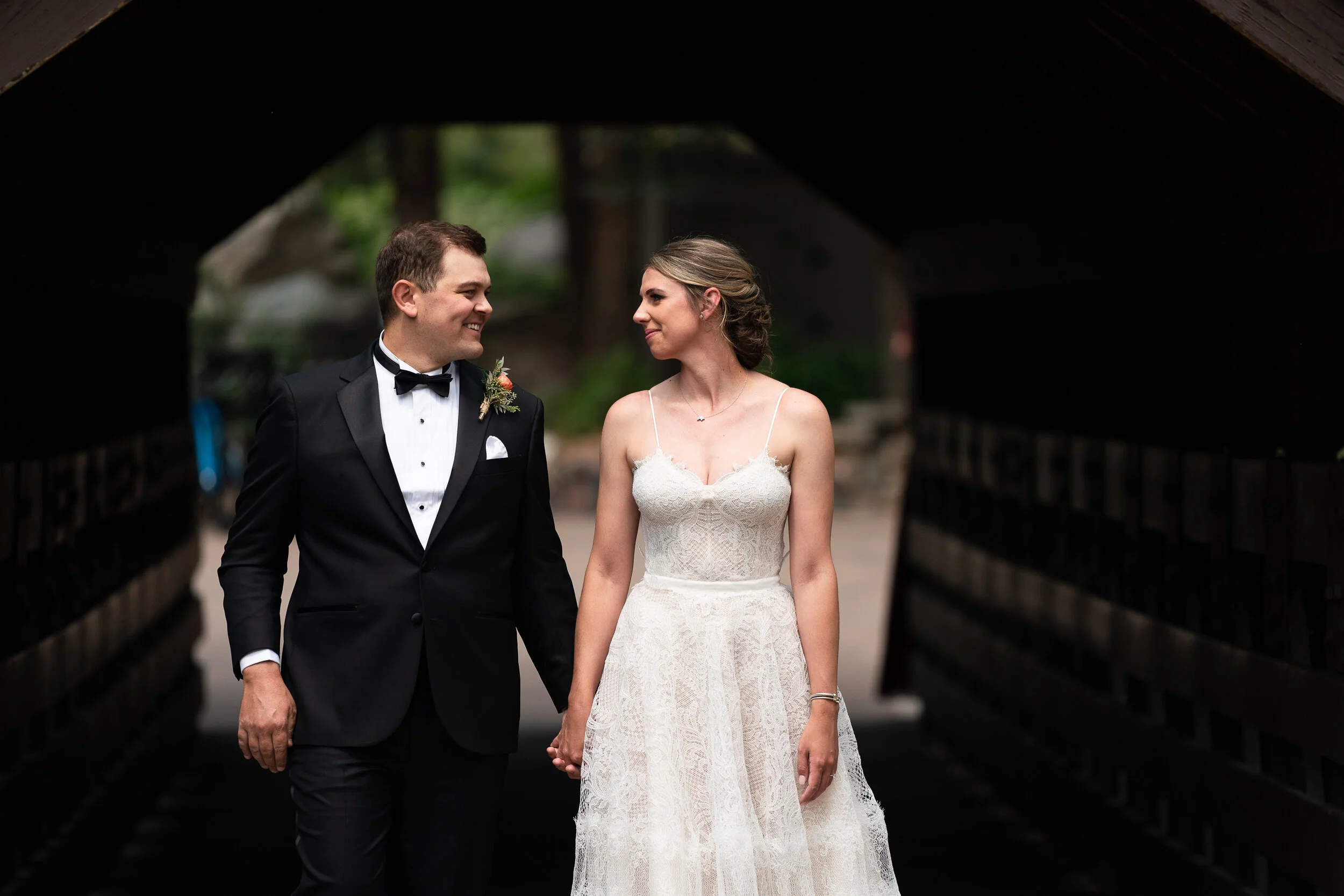 A bride in a white lace gown and a groom in a black tuxedo smiling and holding hands, standing inside a covered bridge.
