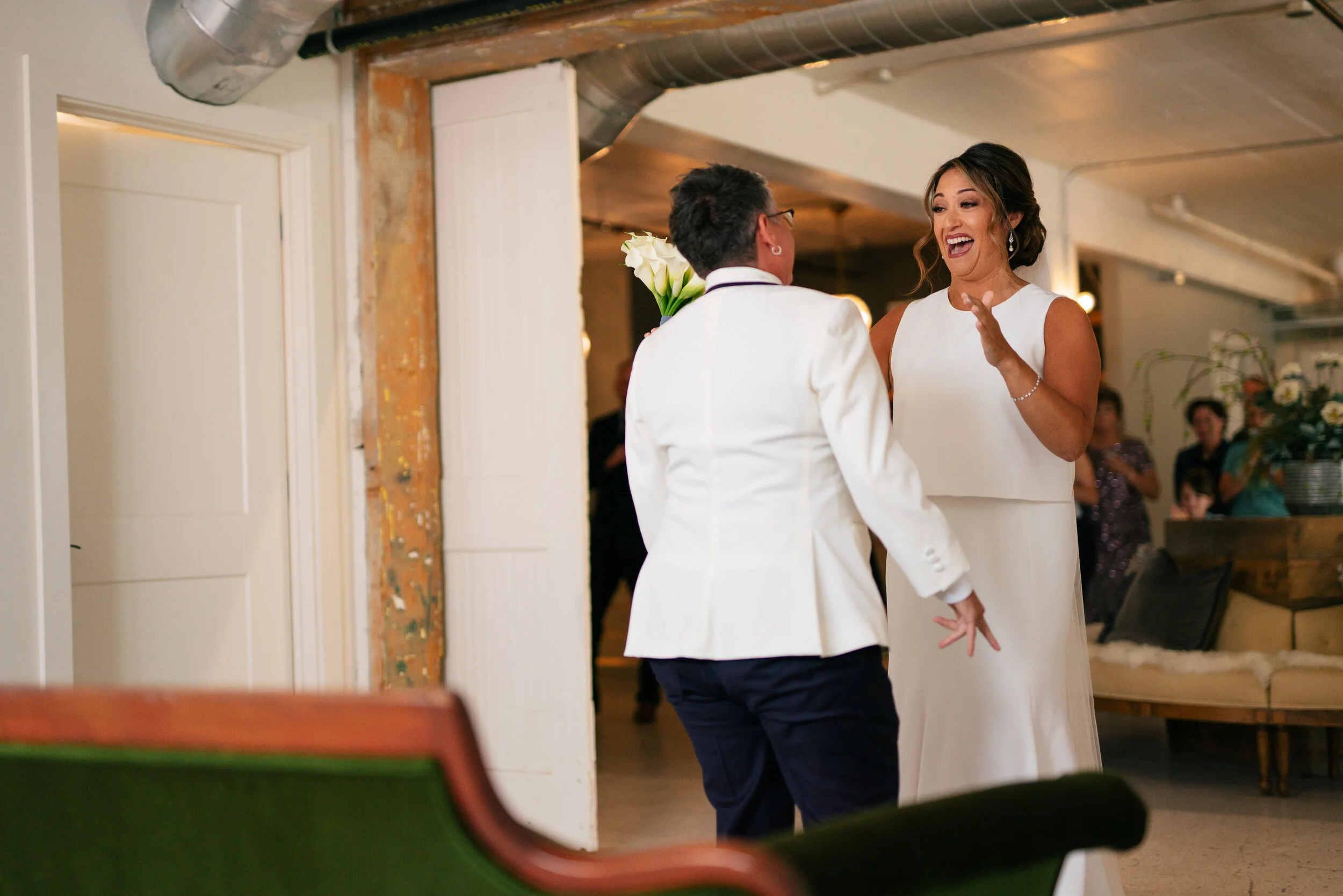 Two people in wedding attire, smiling and interacting indoors with a group of people in the background.
