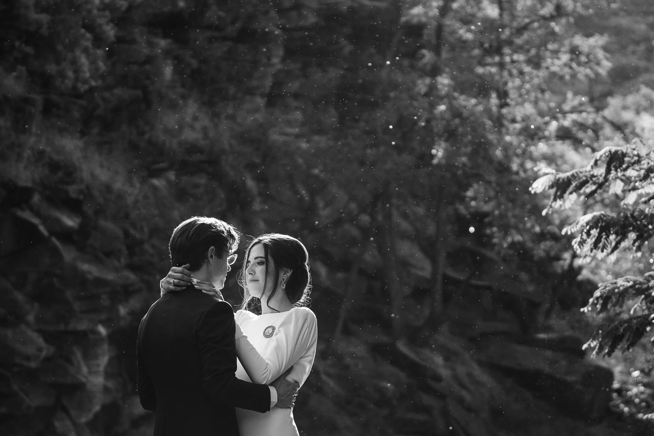 Black and white photo of a couple embracing outdoors with trees in the background.