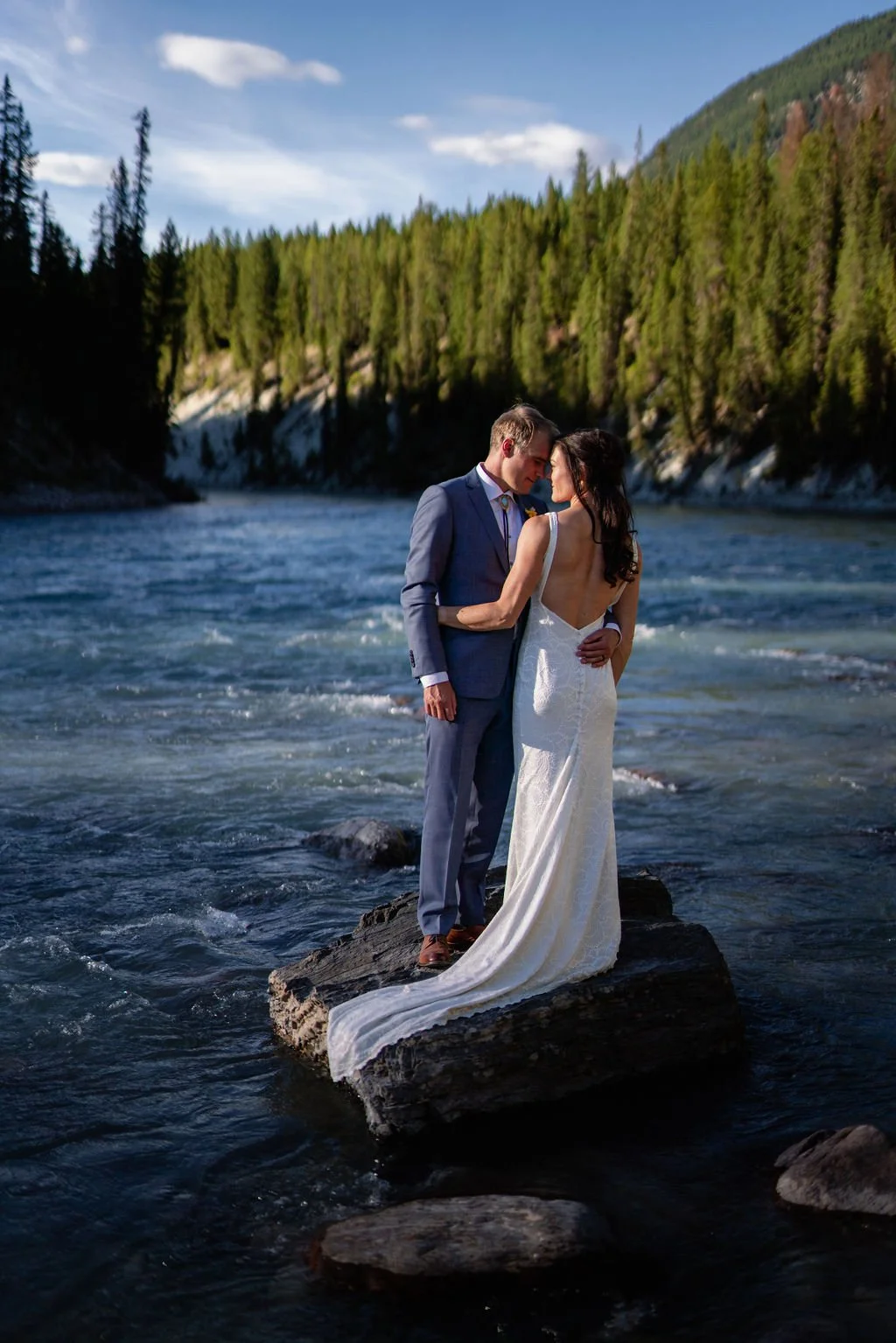 A bride and groom standing on a rock by a river, surrounded by a forest, with the groom in a suit and the bride in a white dress.