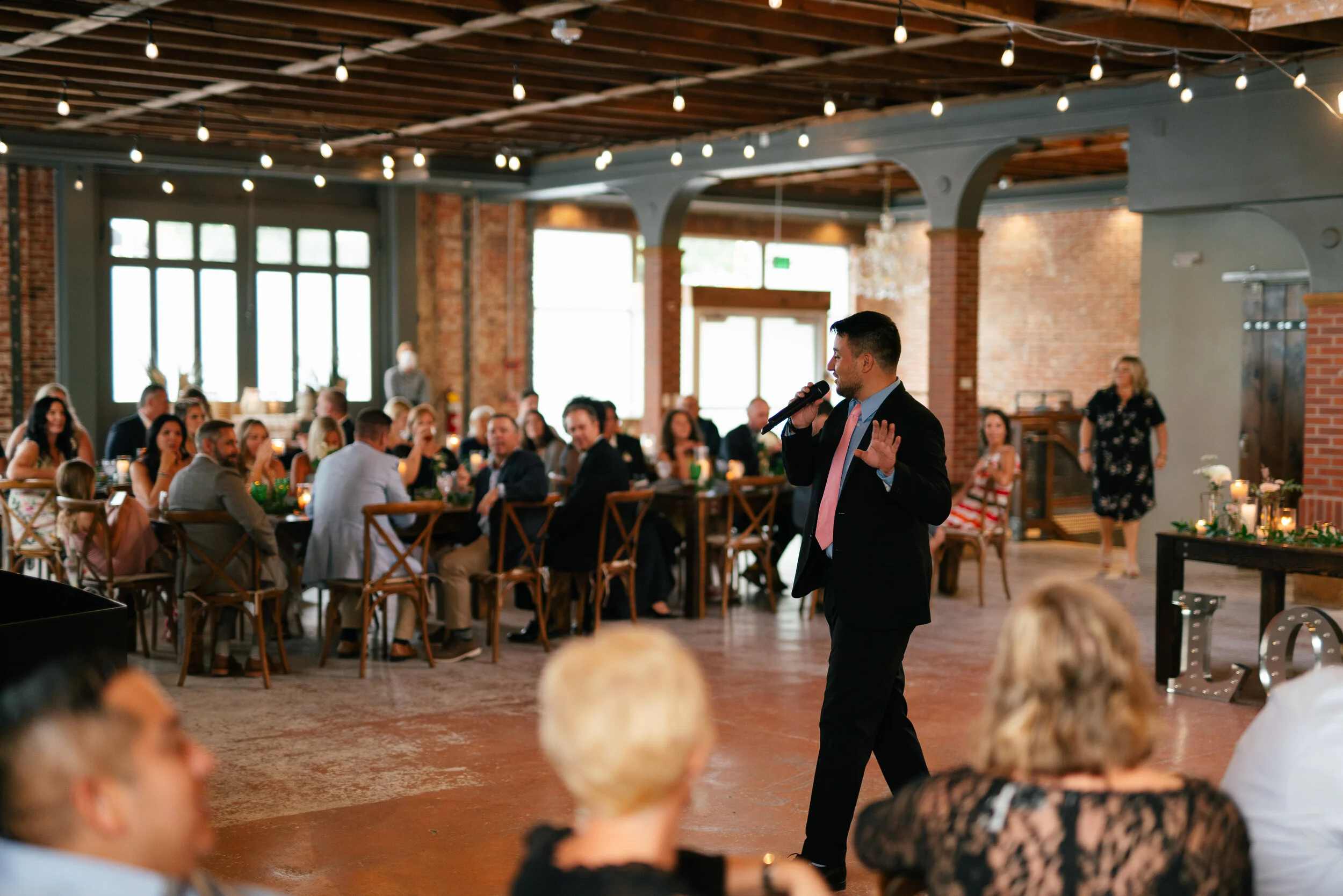 A person in a suit speaking into a microphone in front of seated guests in a restaurant or event space with exposed brick walls and string lights.
