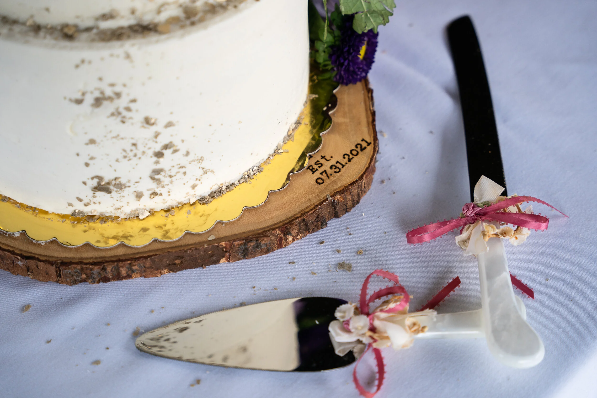 Close-up of a wedding cake on a wooden slab with the date "07.31.2021" engraved. A cake knife and server with ribbon decorations are on the table.