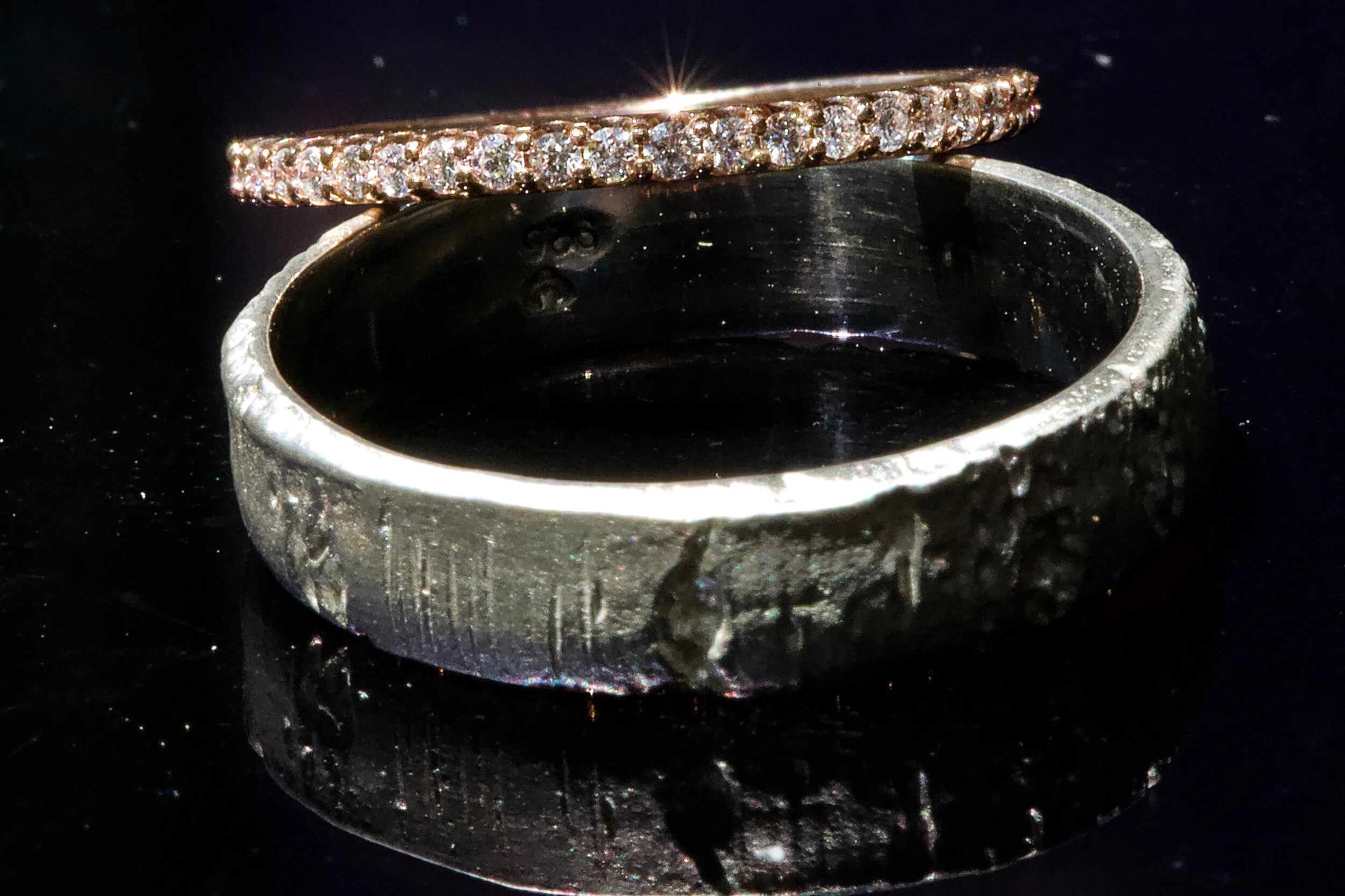 Two rings on a reflective surface; one with small diamonds, the other with a textured silver design.