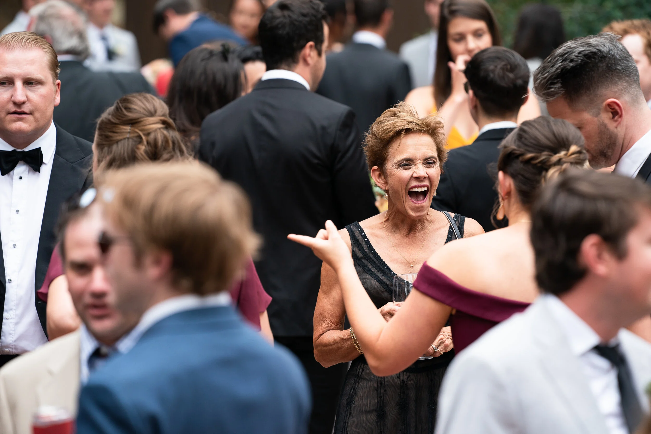 Group of formally dressed people at an outdoor event, with a woman laughing and holding a drink.