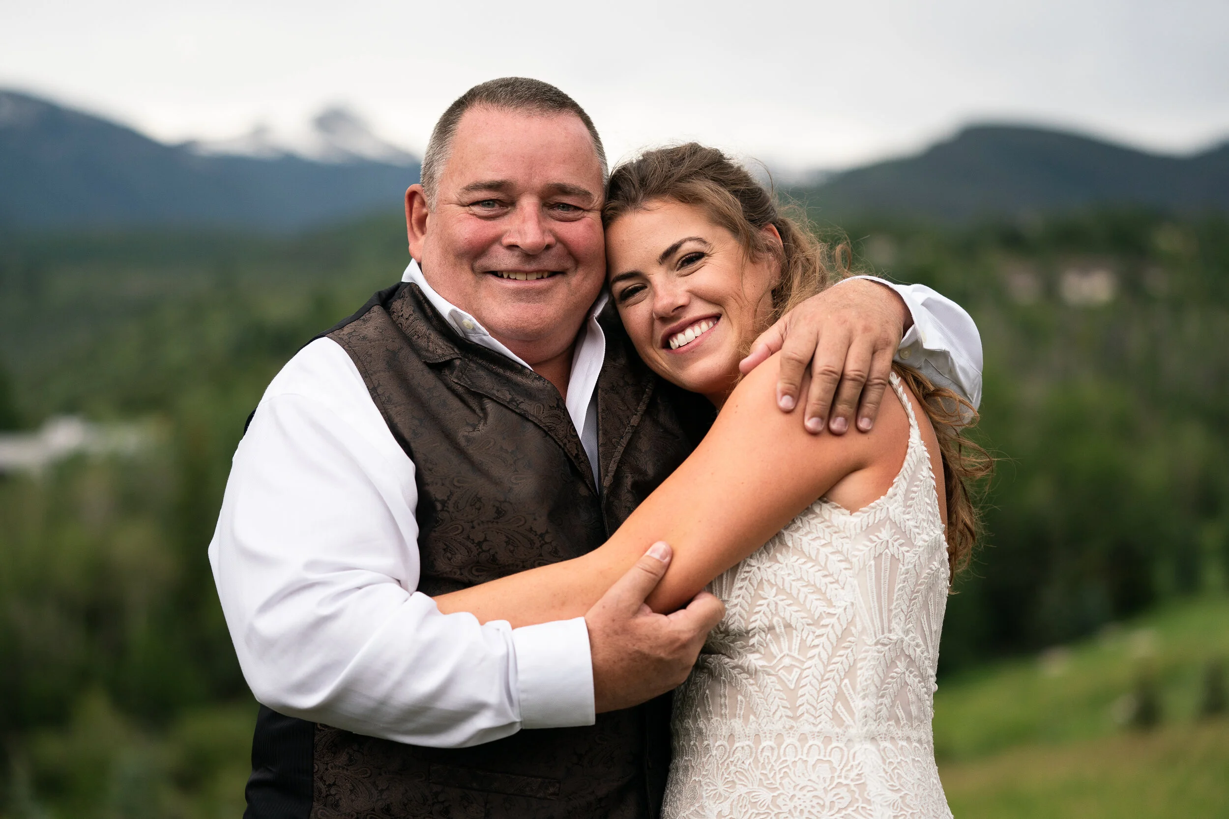 A woman in a white lace dress and a man in a formal vest hug and smile outdoors with a mountainous landscape in the background.