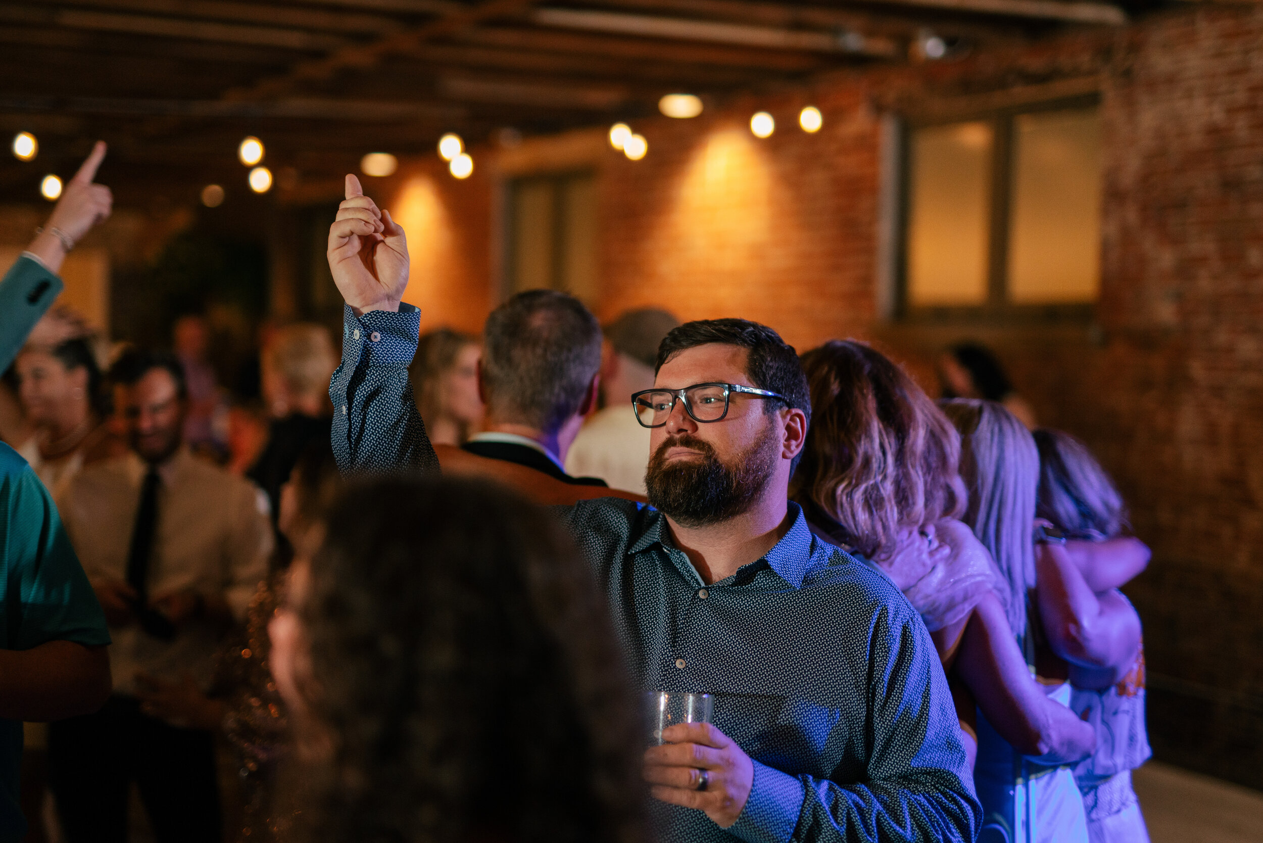 A man with glasses raises his hand at a party, surrounded by blurred people and string lights.