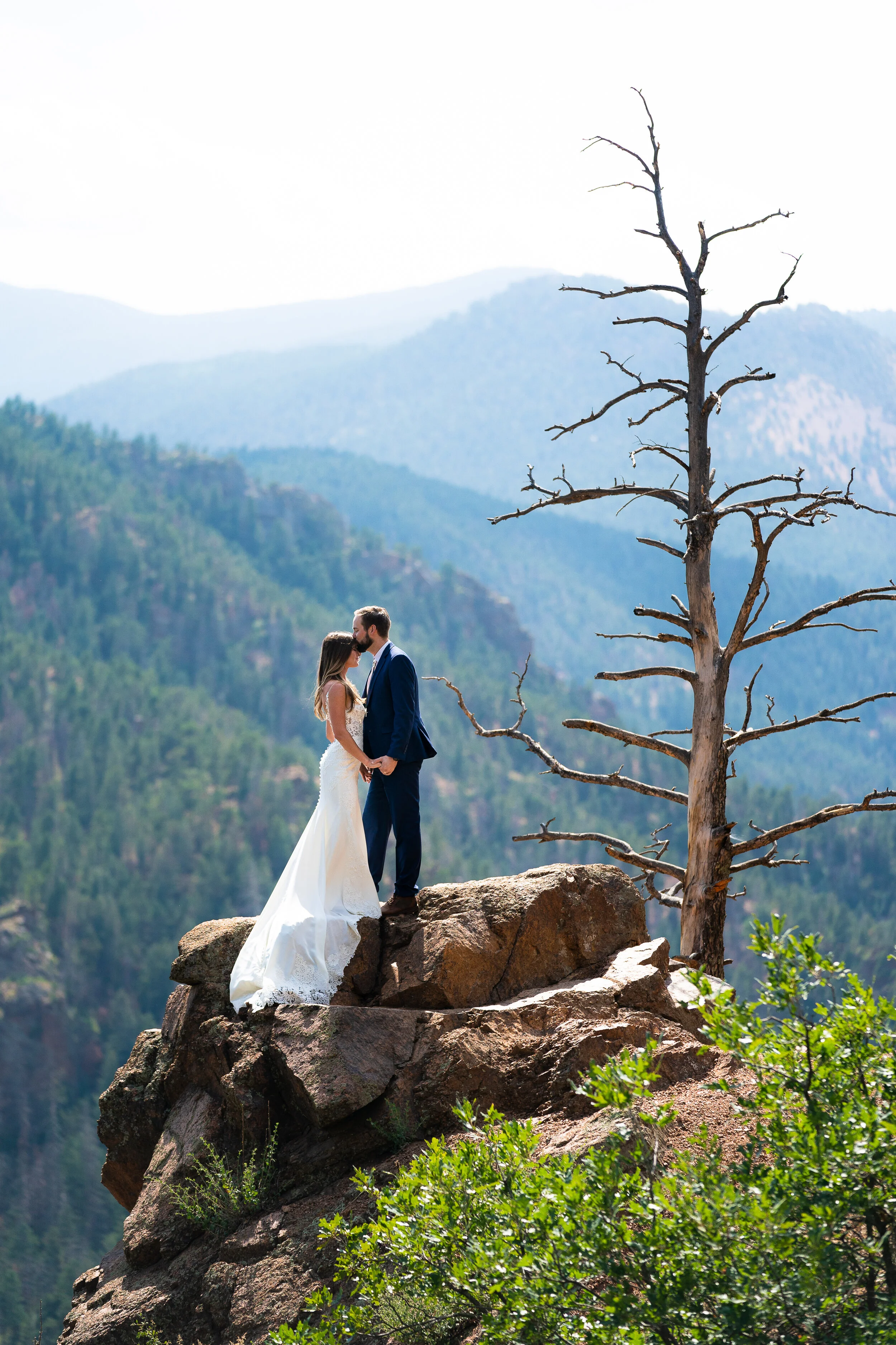 Bride and groom standing on a rocky cliff, surrounded by mountains and trees, sharing a kiss.