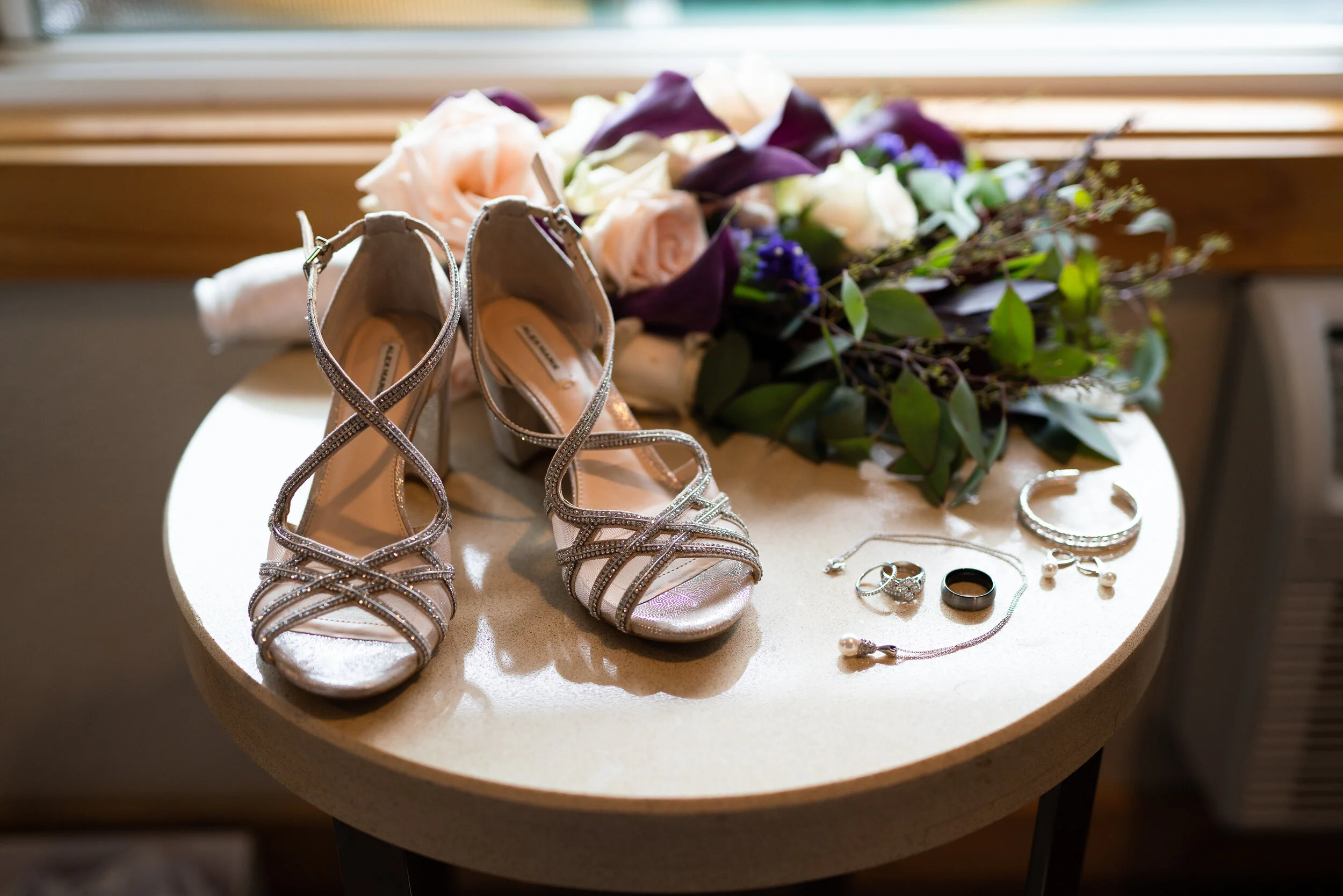 Elegant silver high-heeled shoes and bridal jewelry on a table, with a floral bouquet in the background.