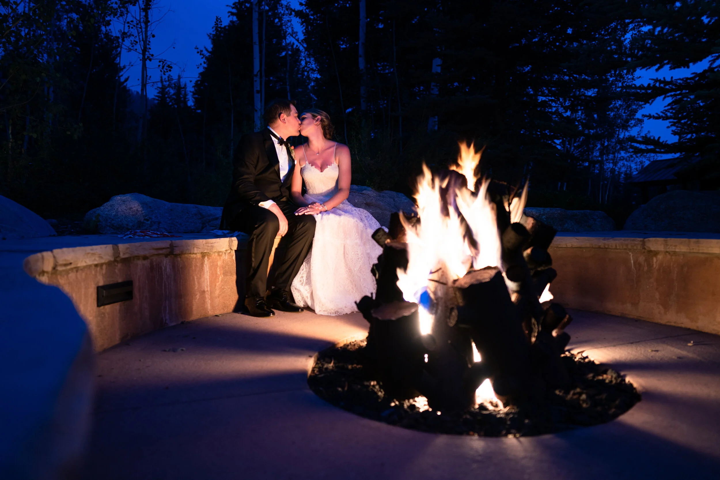 Couple in formal attire kissing by a campfire at night.