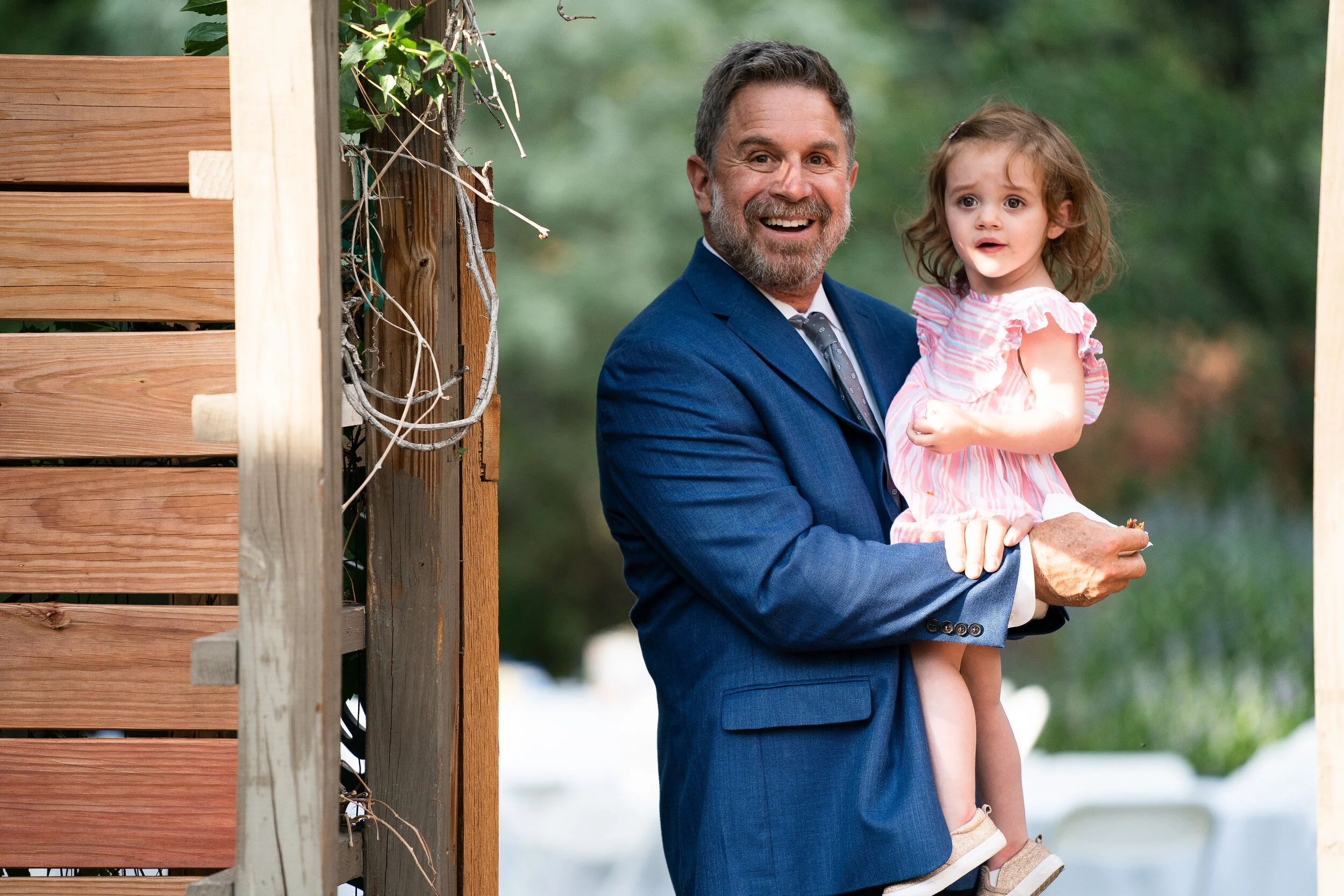 Man in a blue suit holding a young girl wearing a pink dress, standing by a wooden structure outdoors.
