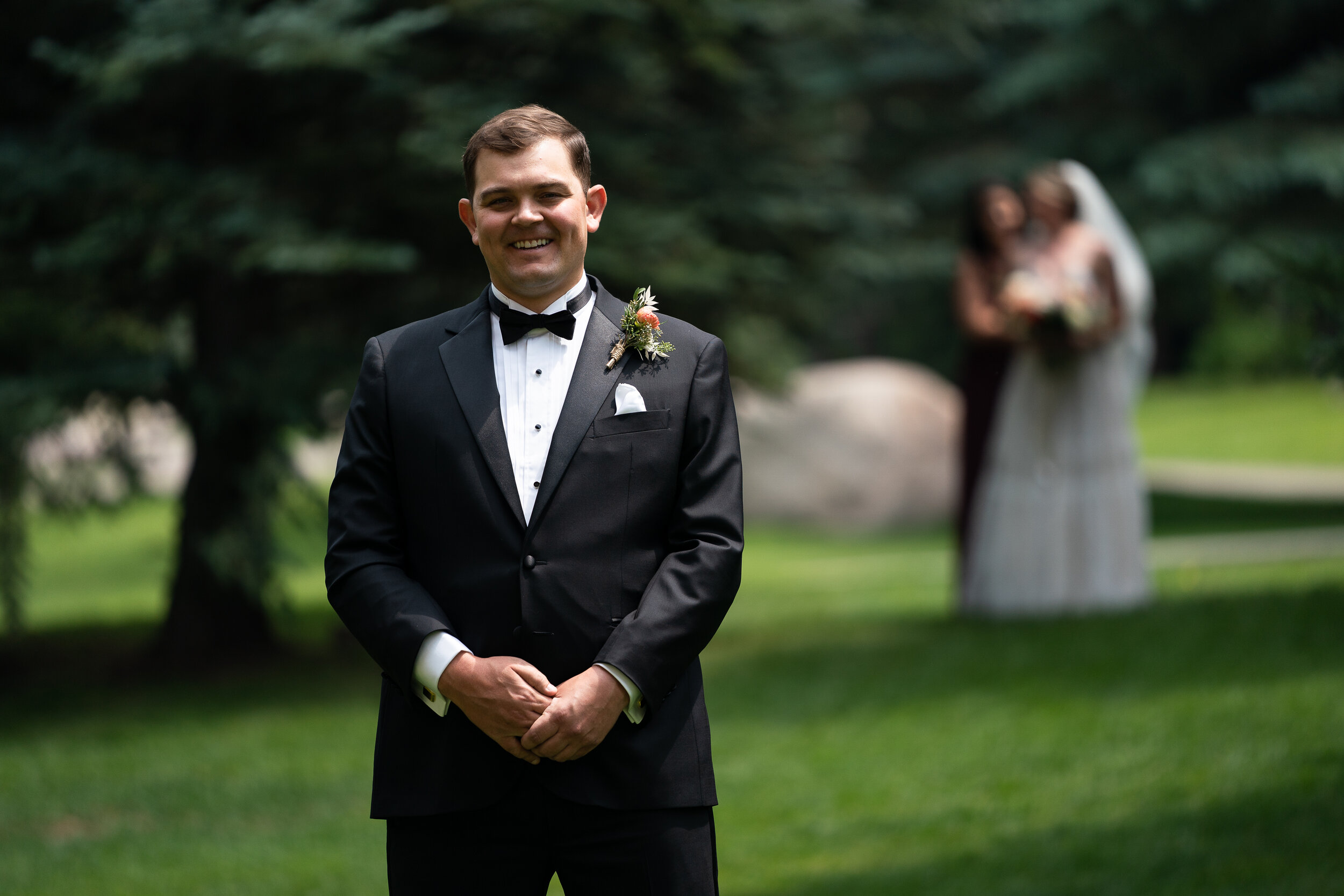 Man in a tuxedo smiling, standing on grass with trees; blurred bride and bridesmaid in the background.