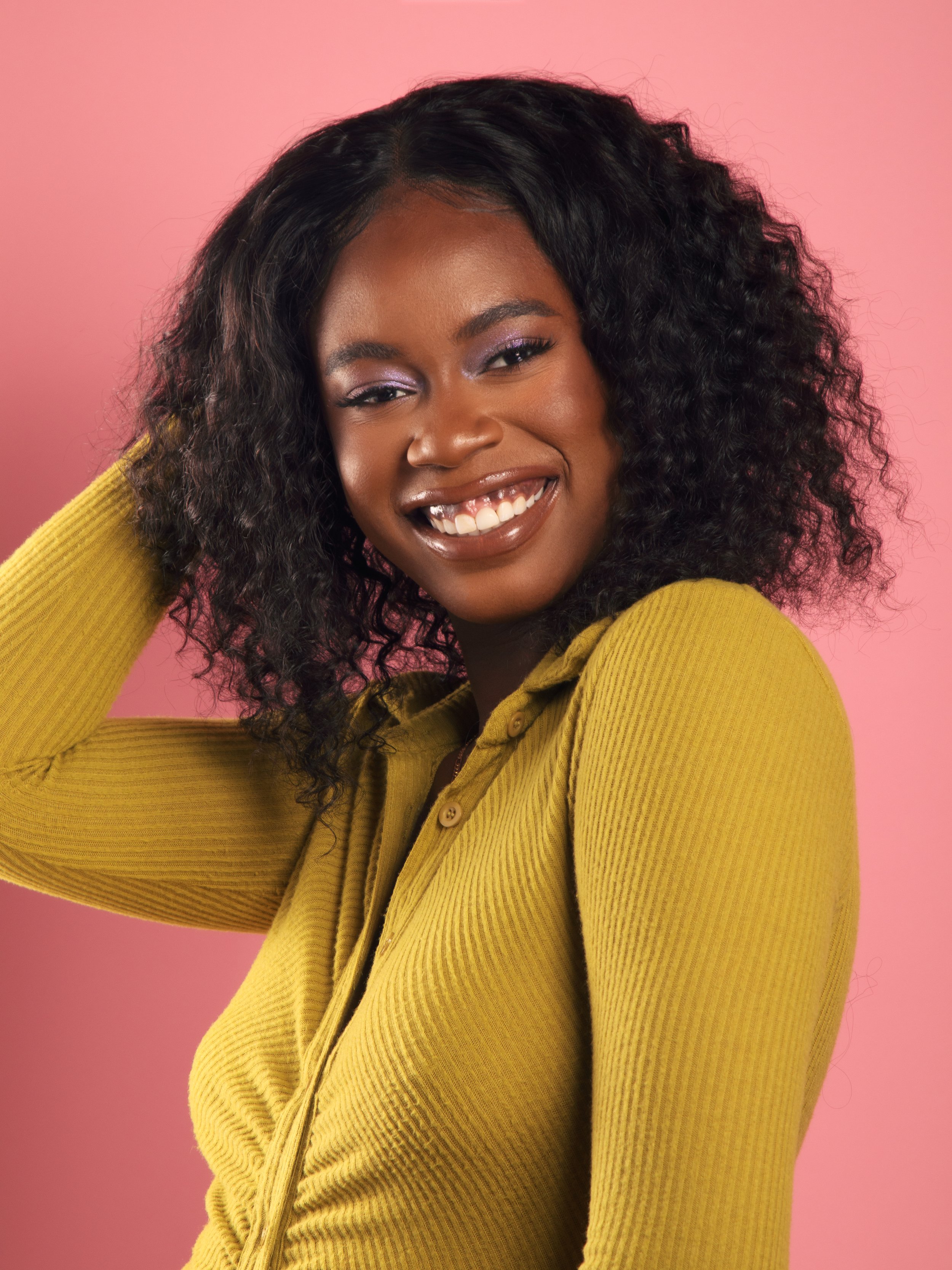 Olivia Bernard is a photographer who is in a green ribbed dress with a pink seamless paper backdrop behind her. She has curly hair that is shourlder length. She is smiling with her top row of teeth showing. She has shimmery purple eyeshadow on.