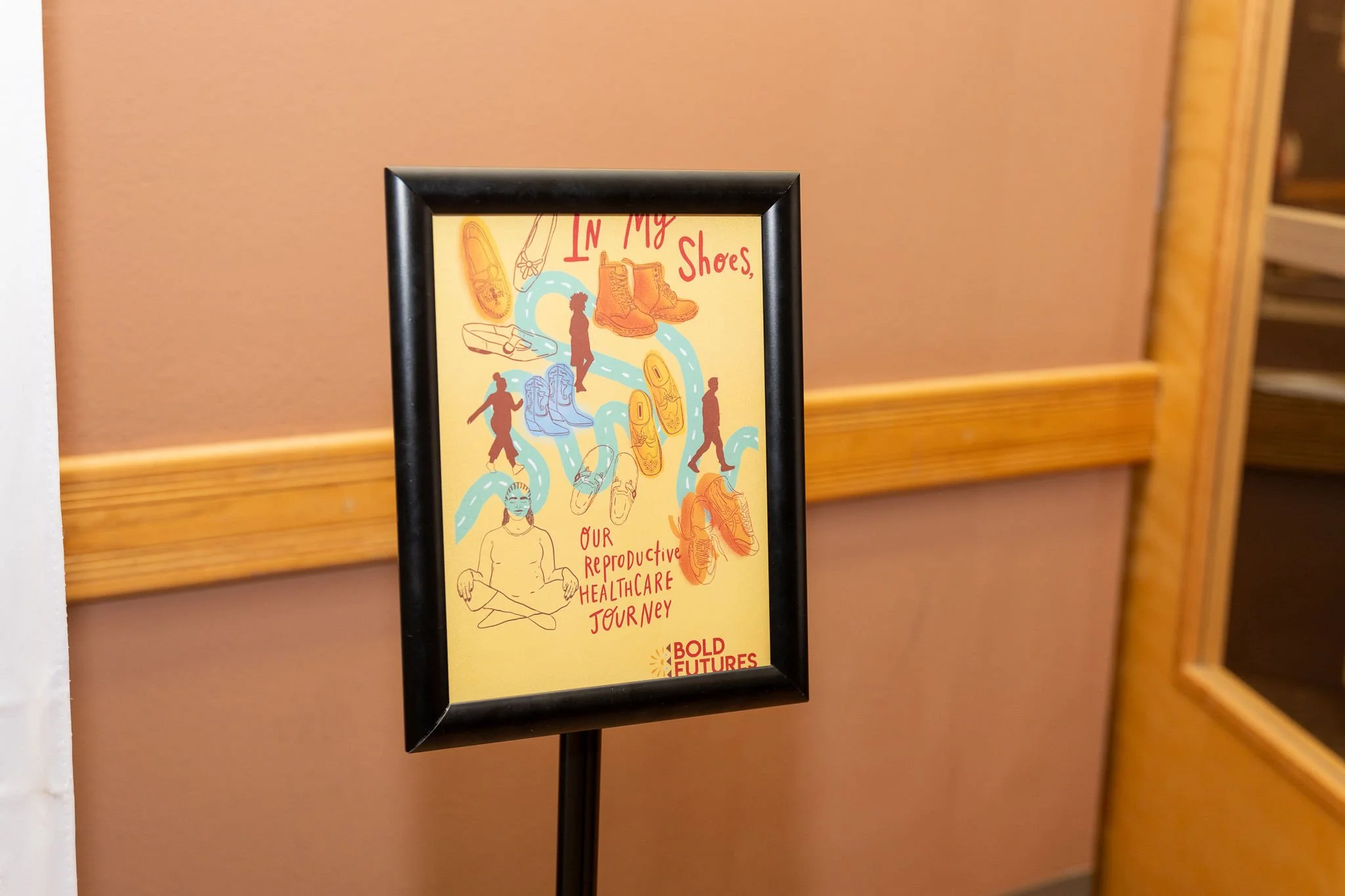 A small poster in a photo frame. The poster has various shoes and human silhouettes, with the text "In my shoes: our productive healthcare journey"