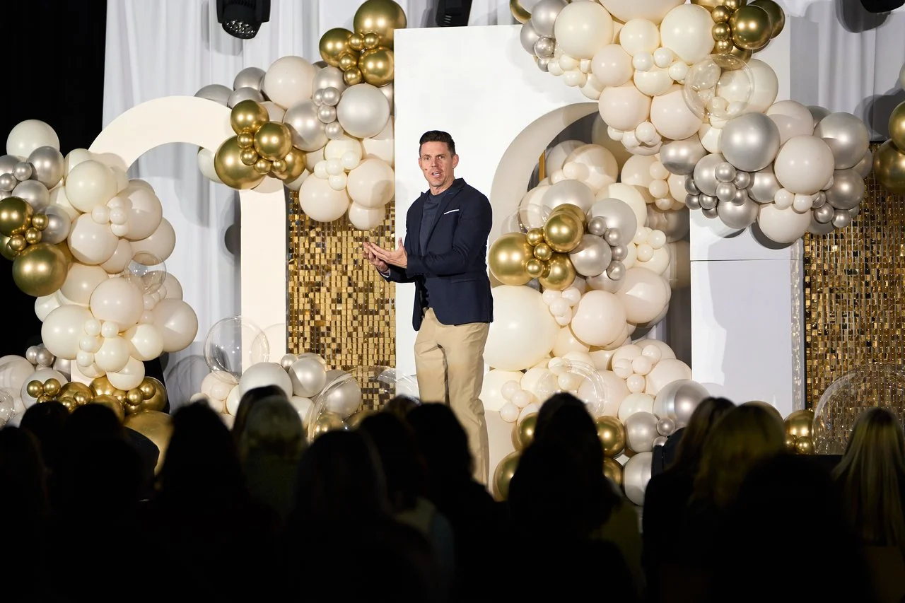 At NuSkin Dream Builders Academy, Ryan Napierski delivers an inspiring speech against a backdrop of grandeur: enormous white arches, a dazzling gold sequin shimmer wall, and opulent balloon clusters in gold, silver, and cream hues. A breathtaking sta